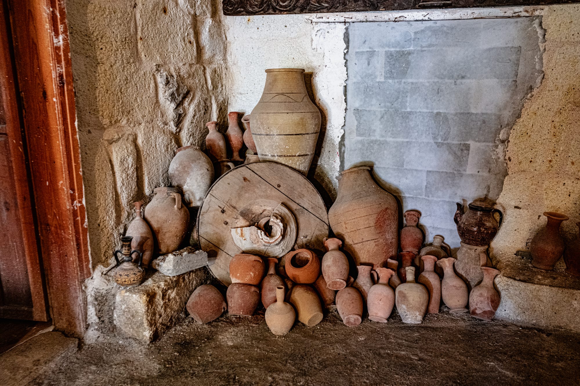 A rustic display of various clay pots and jars at Sultan’s Ceramic in Cappadocia, stacked against a cave wall with a large wooden wheel centerpiece, showcasing the earthy tones and timeless craftsmanship of Anatolian pottery