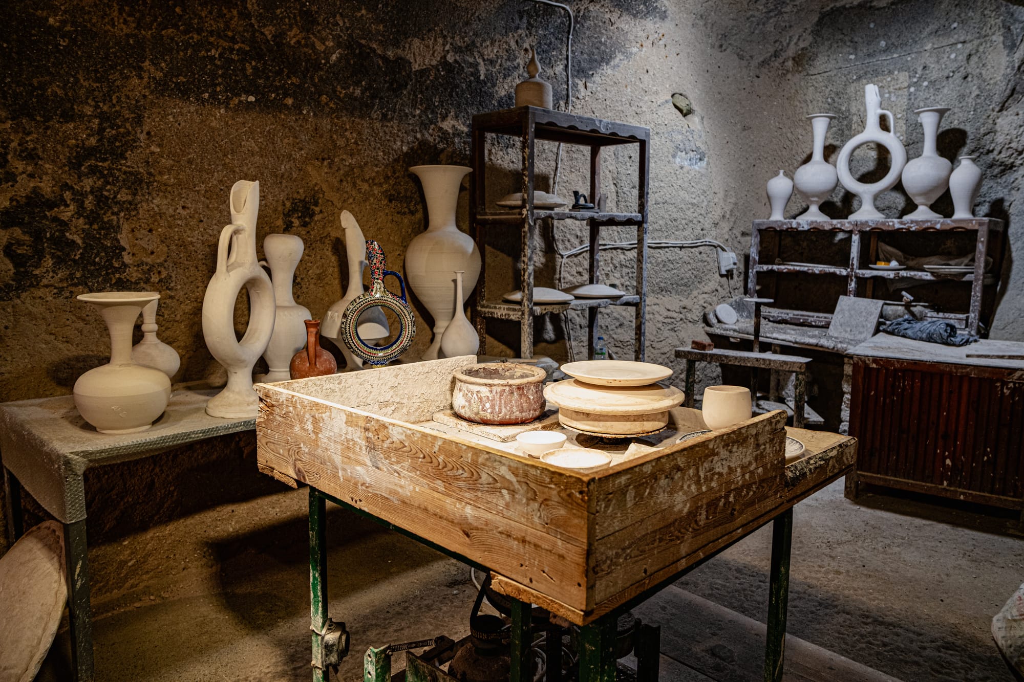 A pottery workshop inside Sultan’s Ceramic in Cappadocia, showing a wooden worktable with a potter’s wheel, shelves of unglazed clay vases, and traditional Cappadocian pottery in various stages of completion