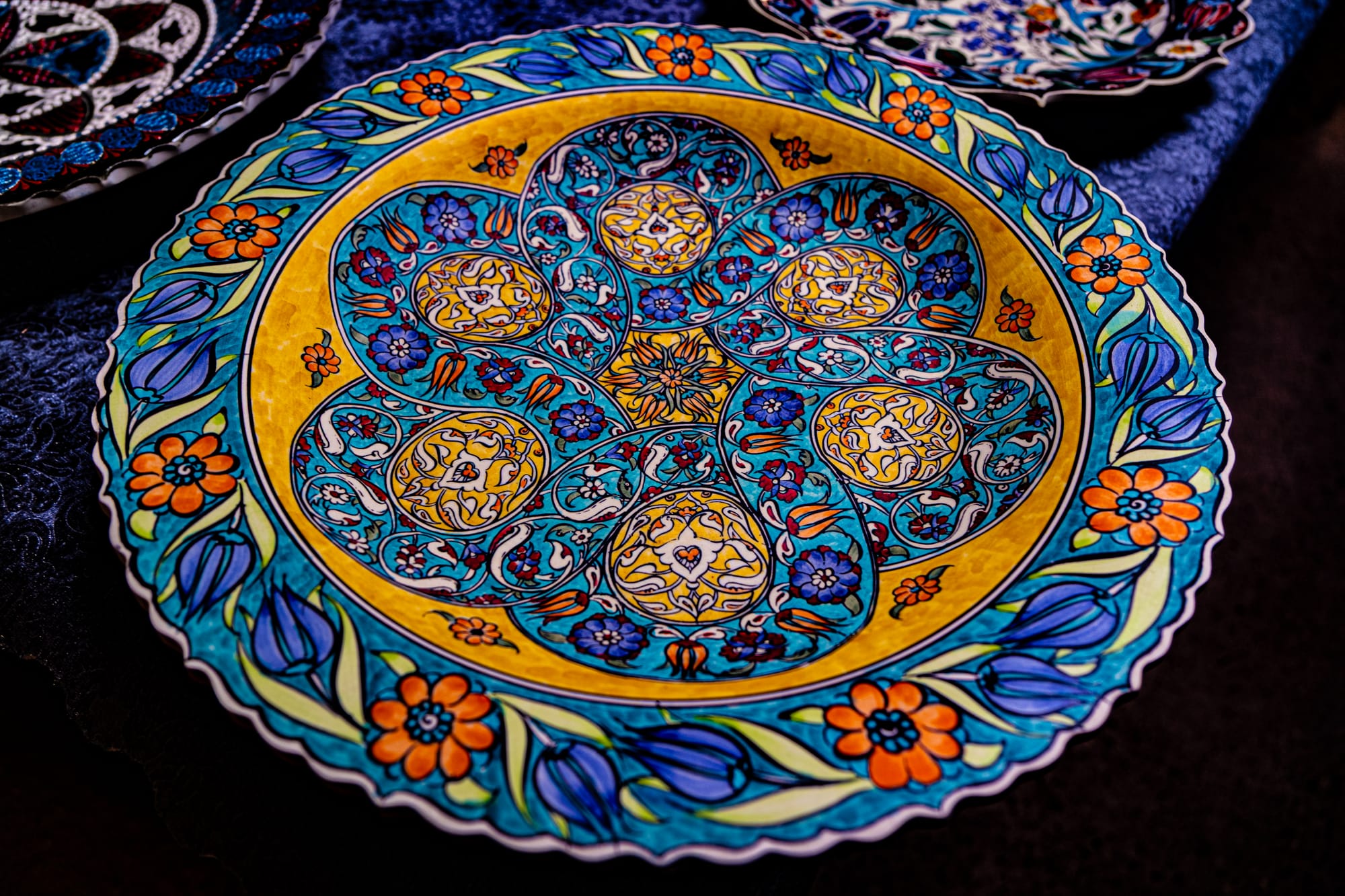 A vibrantly painted ceramic plate from Sultan’s Ceramic in Cappadocia, featuring an intricate floral and geometric design in vivid blue, yellow, orange, and turquoise hues, with delicate hand-painted patterns and a scalloped edge