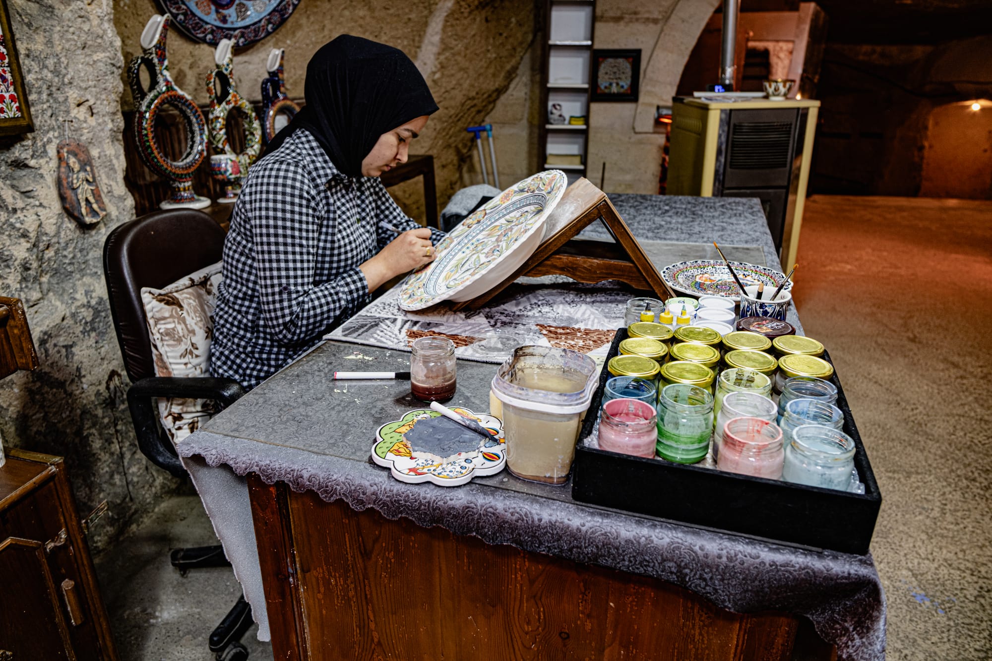 A female artisan at Sultan’s Ceramic in Cappadocia, wearing a black headscarf and checkered shirt, sits at a worktable meticulously painting detailed patterns onto a large ceramic plate, with an array of colorful paint jars and brushes laid out beside her