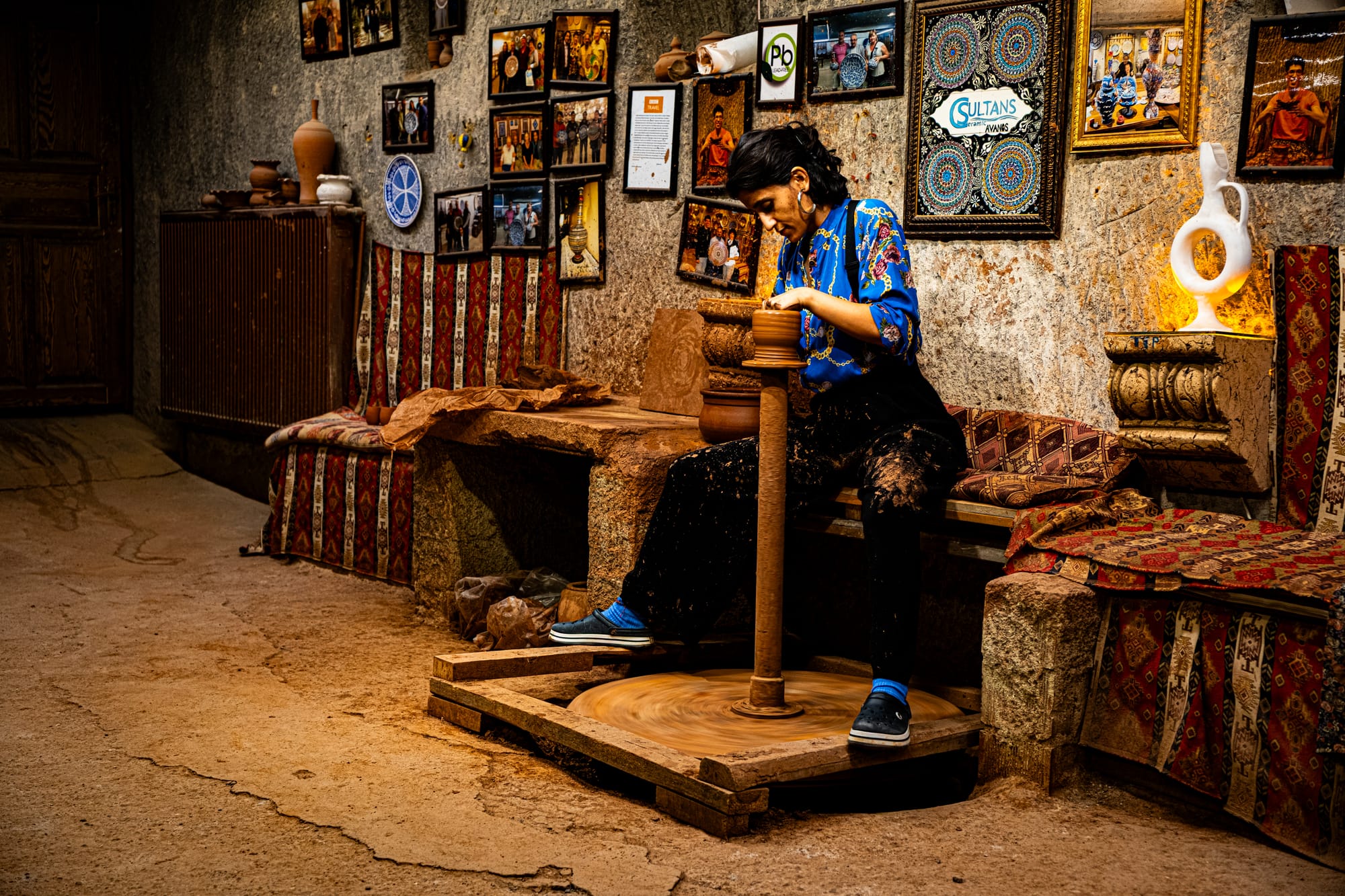 A female artisan at Sultan’s Ceramic in Cappadocia works intently at a traditional potter’s wheel inside a cave workshop, surrounded by framed photos, pottery, and patterned textiles
