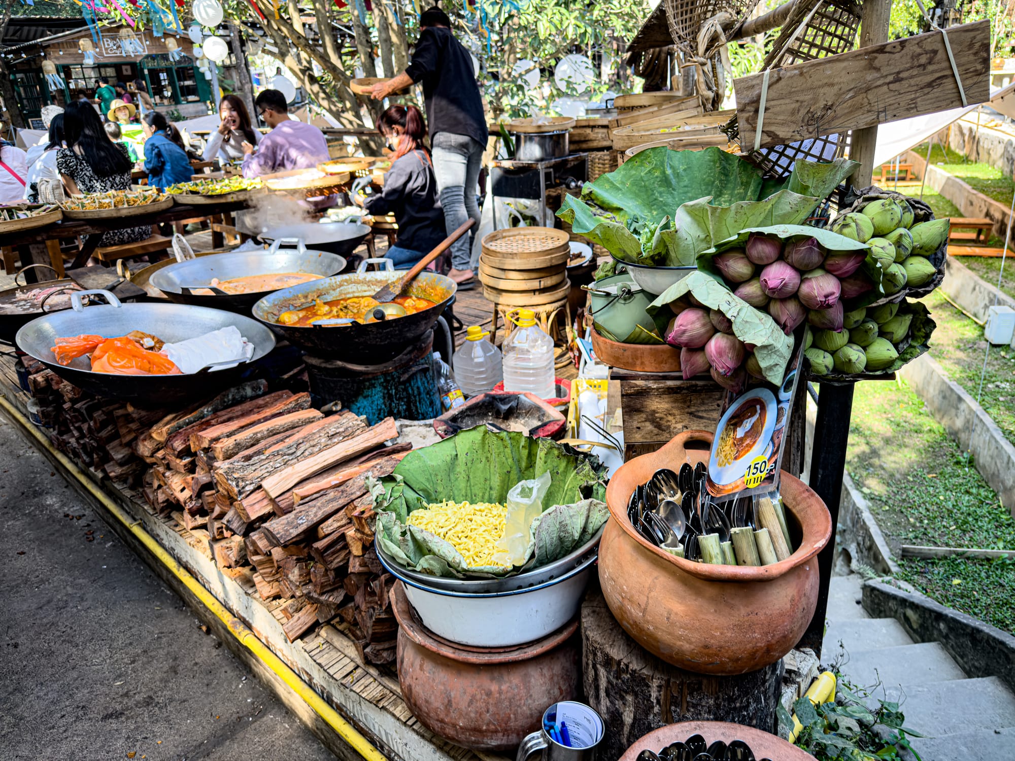 Outdoor food stall at Baan Kang Wat showing woks of curry over wood fire, baskets of noodles wrapped in banana leaves, lotus flowers, and clay pots filled with utensils, with people dining in the background