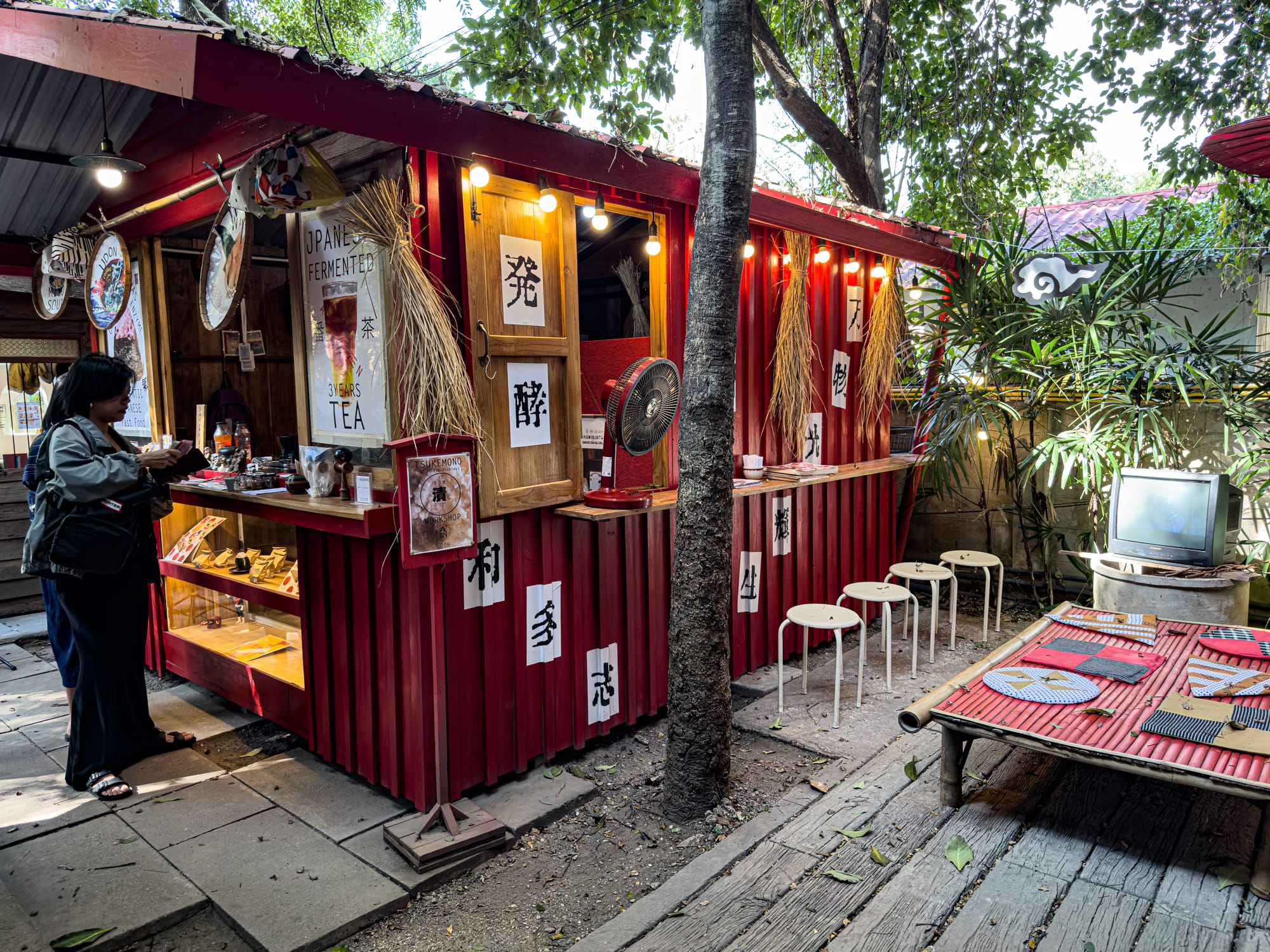 Small red tea shop at Baan Kang Wat in Chiang Mai, featuring Japanese fermented tea, handwritten Japanese signs, bamboo accents, stools lined up at the counter, and a rustic outdoor seating setup