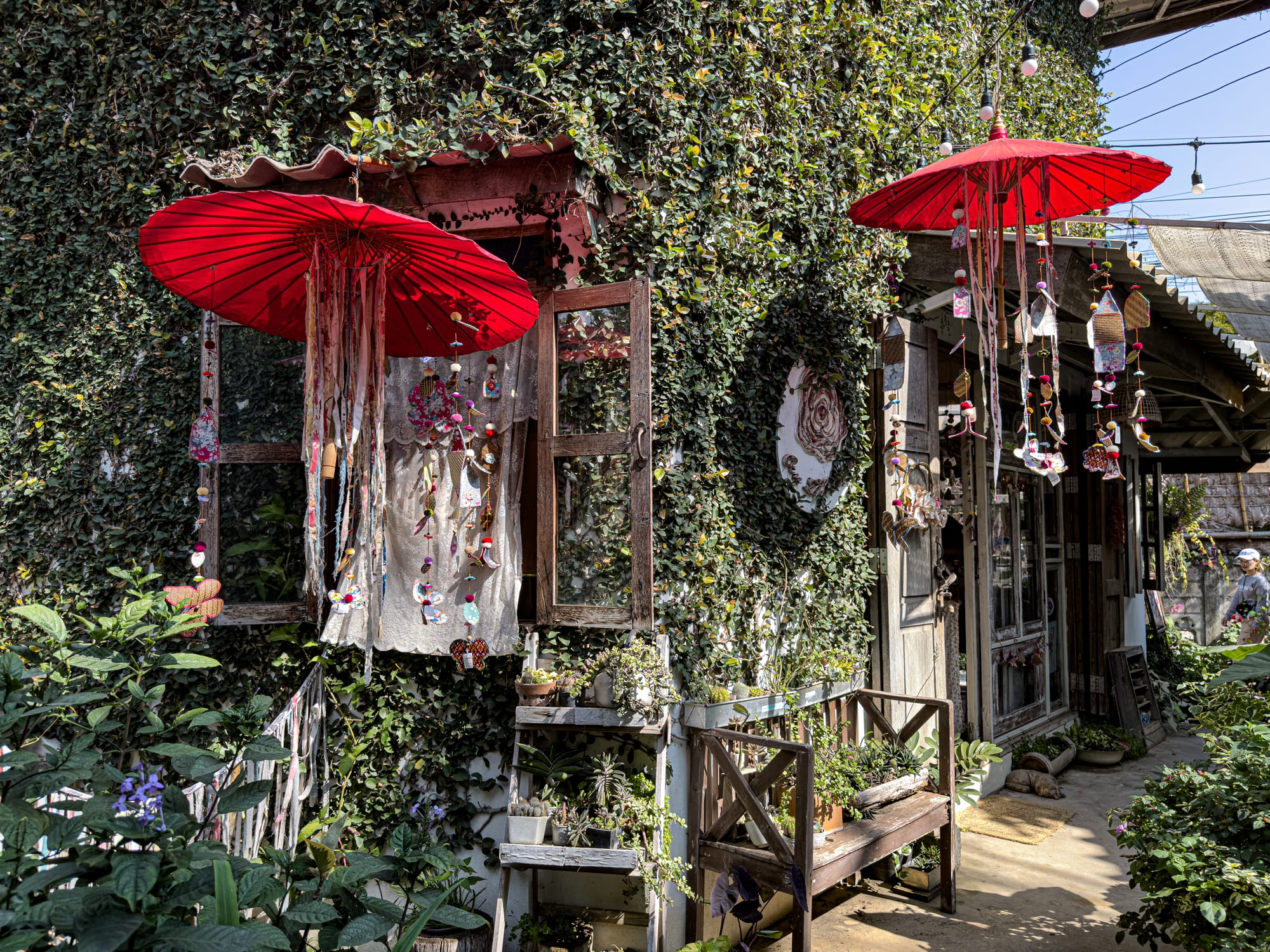 Exterior of a shop at Baan Kang Wat with red umbrellas, dangling handmade decorations, and lush greenery covering the walls