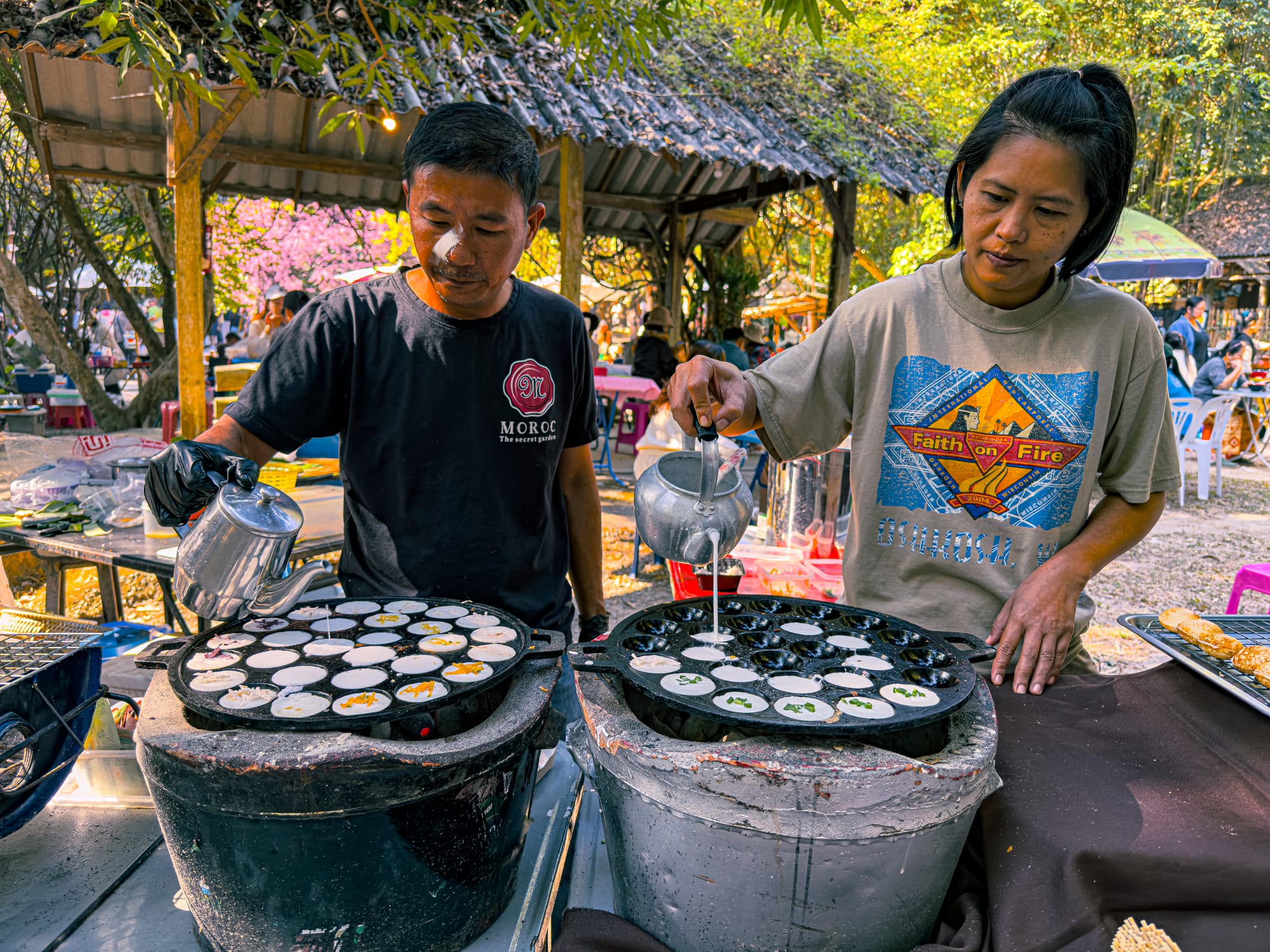 Two street food vendors at Bamboo Saturday Market in Chiang Mai pour coconut milk batter into traditional cast iron pans over charcoal stoves to make fresh khanom krok, a Thai coconut pancake, surrounded by a lively outdoor setting
