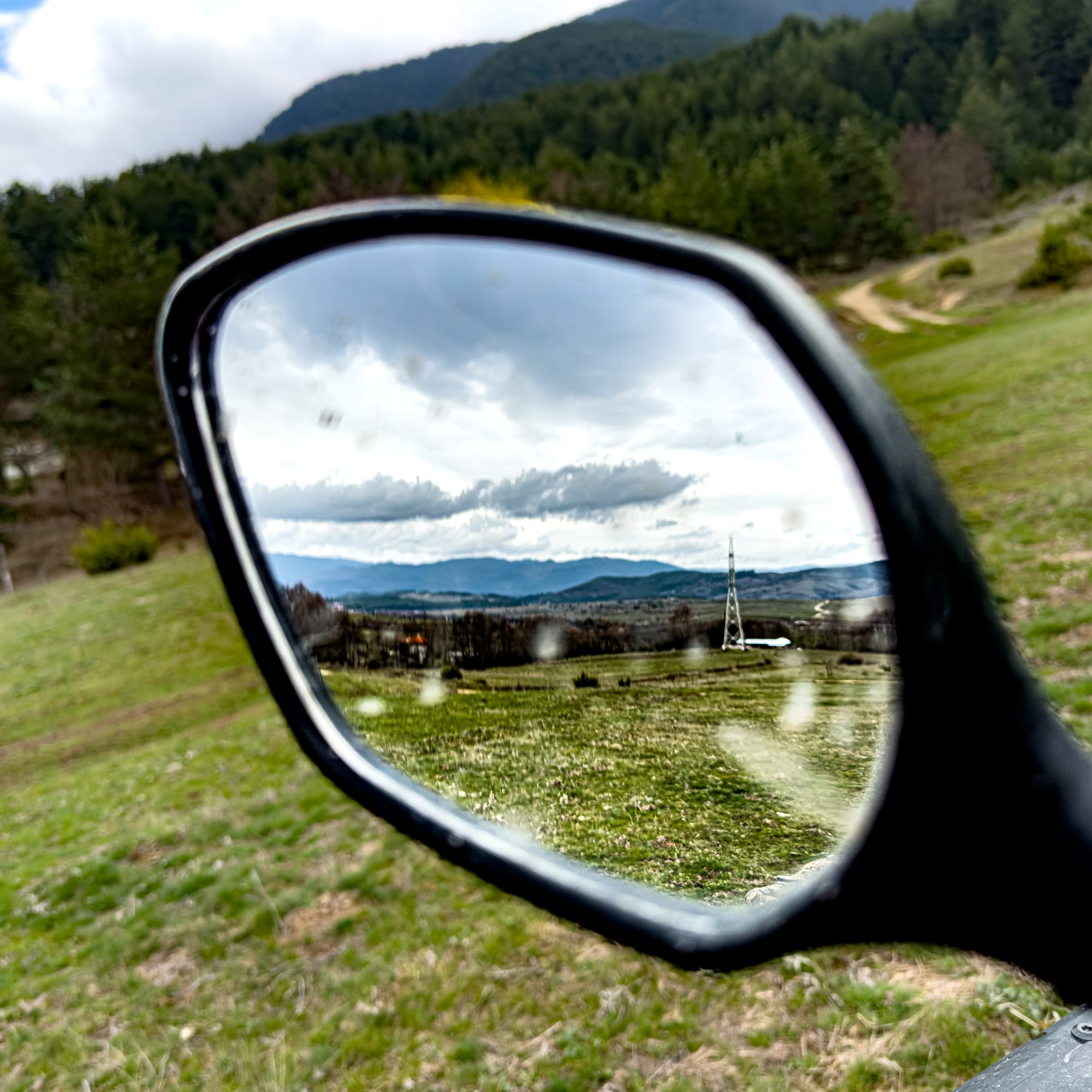 Close-up of an ATV side mirror reflecting distant mountains, grassy fields, and a cloudy sky, with forested slopes in the background near Bansko, Bulgaria