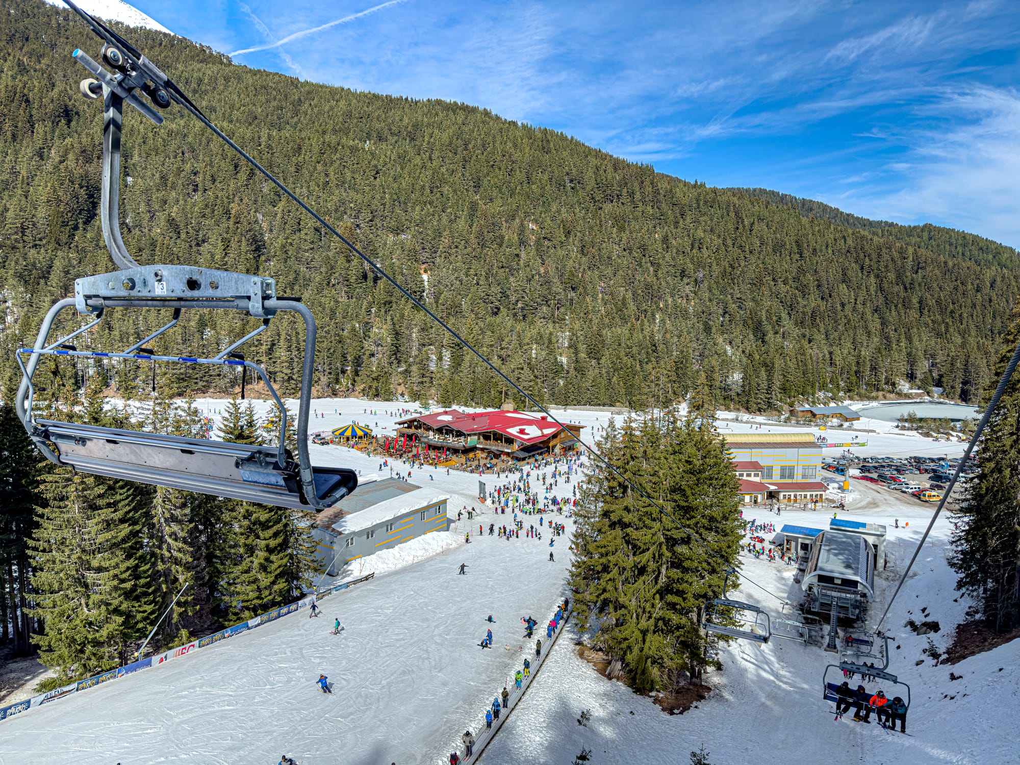 View from a chairlift in Bansko’s Pirin Mountains, looking down at the ski base area with skiers on the snow, a busy lodge with a red roof, and surrounding pine forests