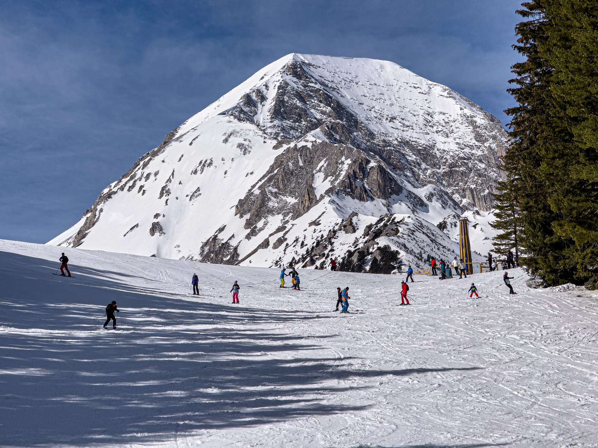 Group of skiers on a snowy slope in Bansko, Bulgaria, with a towering snow-covered mountain peak behind them under a clear blue sky