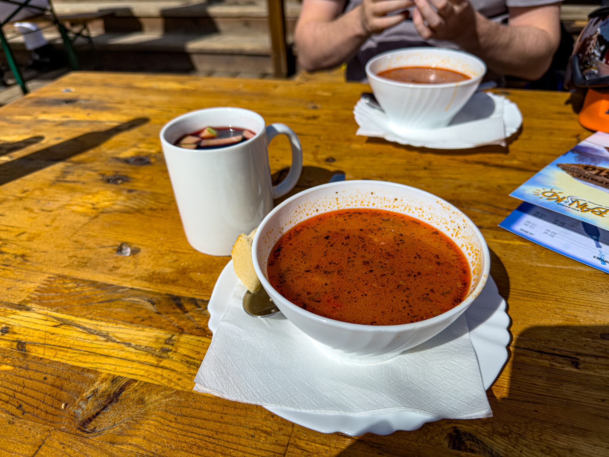 Close-up of a wooden table with a white bowl of steaming red soup and a mug of mulled wine with fruit pieces, served outdoors at a ski resort in Bansko’s Pirin Mountains
