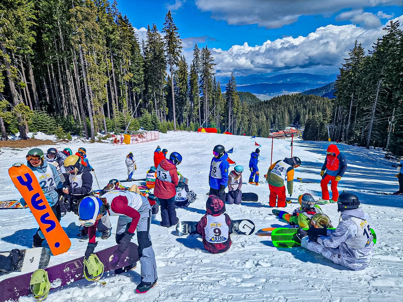 Group of snowboarders wearing helmets and numbered vests preparing their boards on a snowy slope in Bansko’s Pirin Mountains, surrounded by tall pine trees with mountain views in the distance