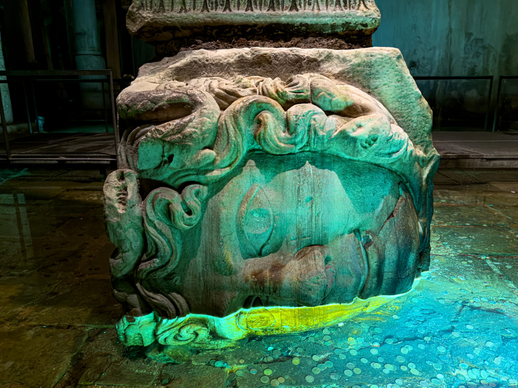 Close-up of a sideways Medusa head sculpture at the base of a column in the Basilica Cistern, dimly lit with green and blue lights and surrounded by coins on the wet stone floor
