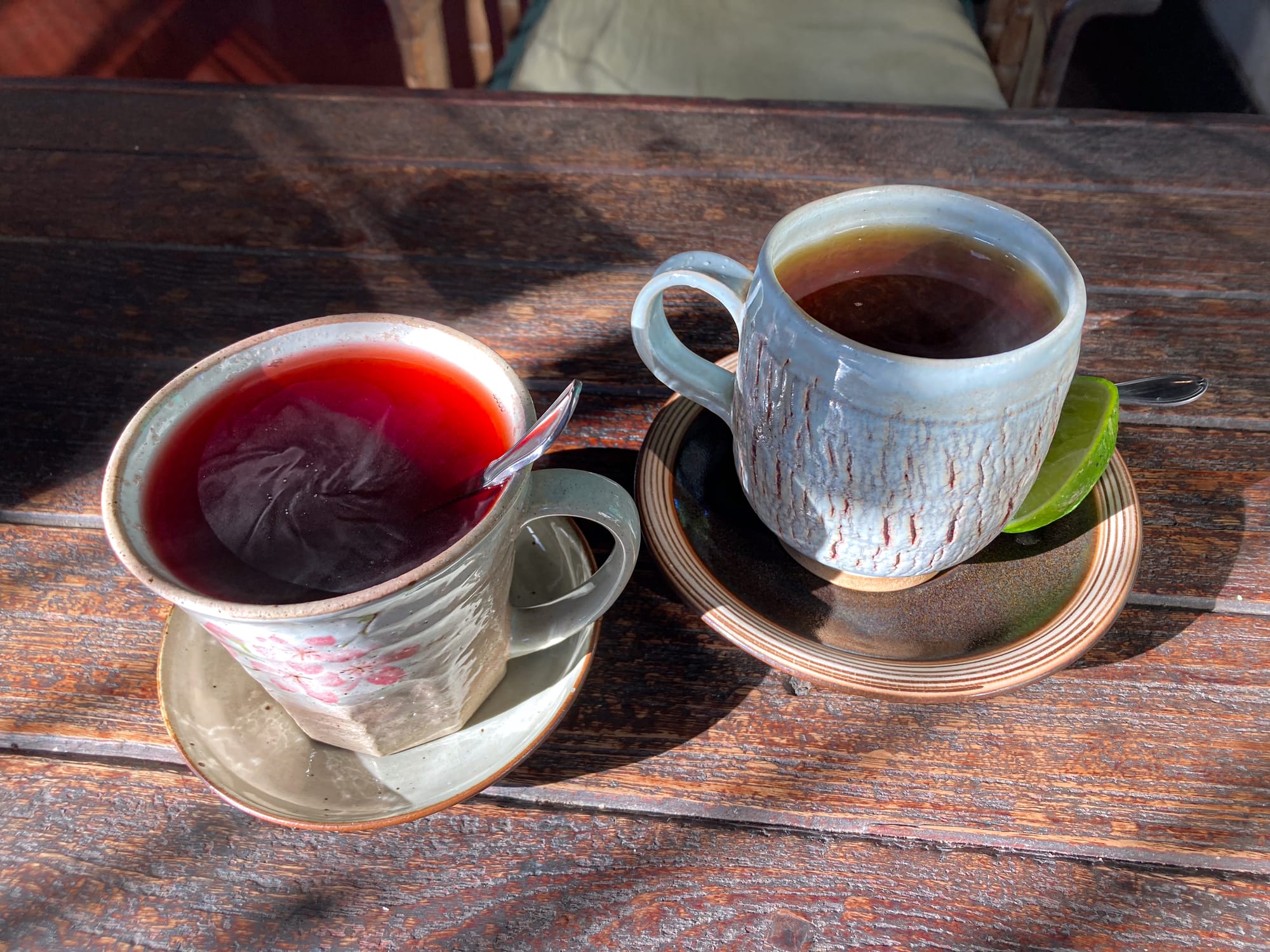Two cups of tea at Bird’s Nest Café in Chiang Mai, one deep red herbal tea and one black tea with a slice of lime on the saucer, both served in rustic handmade ceramic mugs on a wooden table