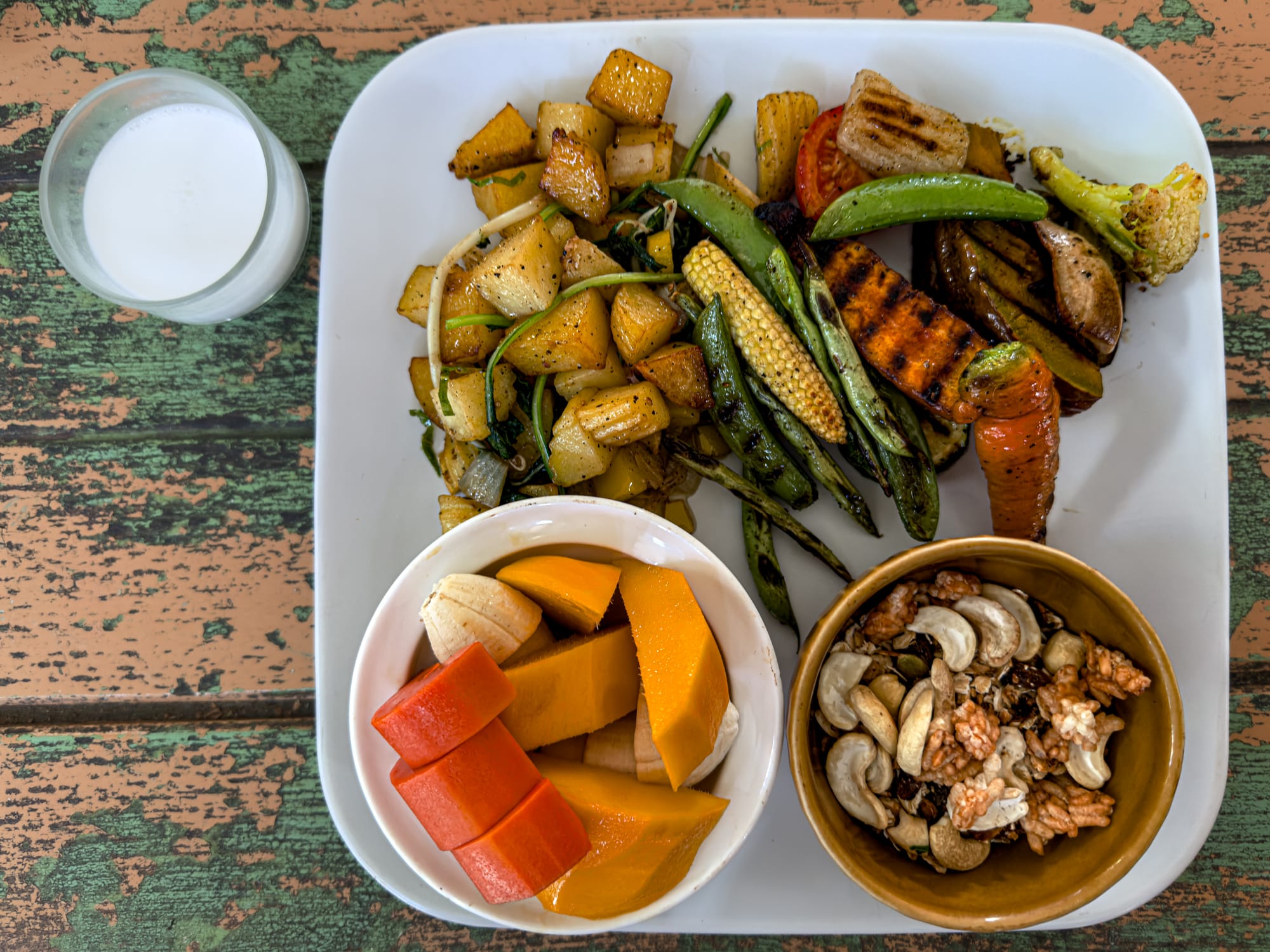 Colorful breakfast plate at Bird’s Nest Café in Chiang Mai featuring grilled vegetables, sautéed potatoes, fresh papaya, banana, and mango, alongside a bowl of granola with cashews and walnuts, served with a glass of coconut milk