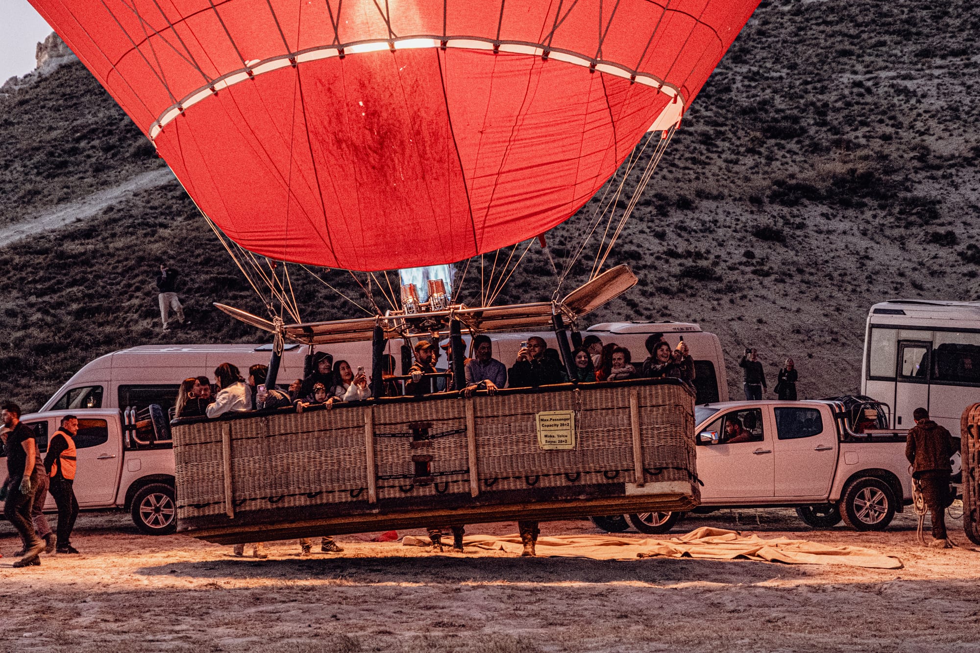 A group of people stand inside a wicker hot air balloon basket as the flame ignites under a large red balloon canopy, preparing for takeoff at dawn in Cappadocia, with support vehicles and crew surrounding it on the ground