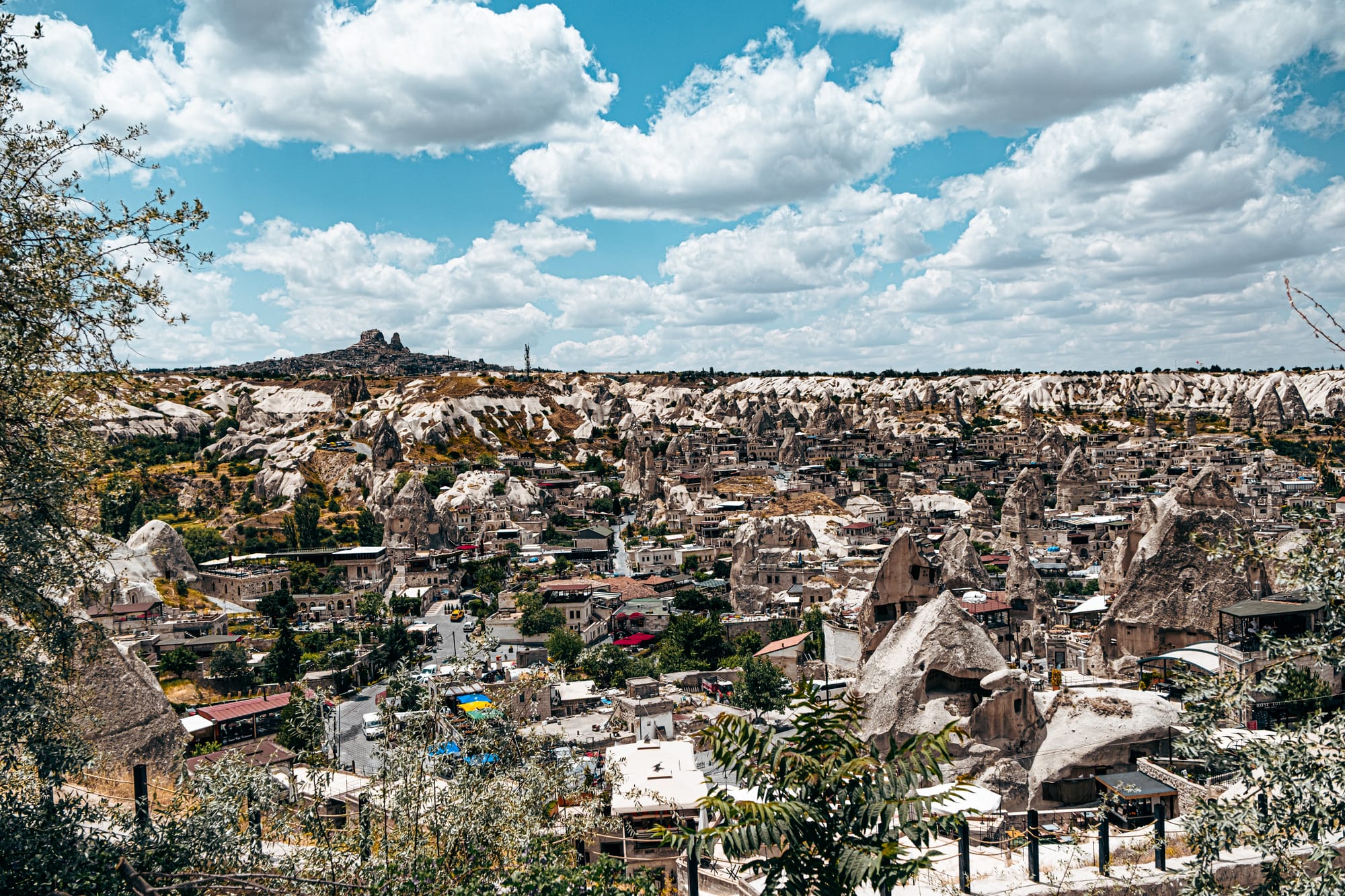 Panoramic view of Göreme in Cappadocia, Turkey, with stone houses and fairy chimneys spread across the valley, framed by rugged white cliffs and Uçhisar Castle rising in the distance under a bright blue sky with scattered clouds
