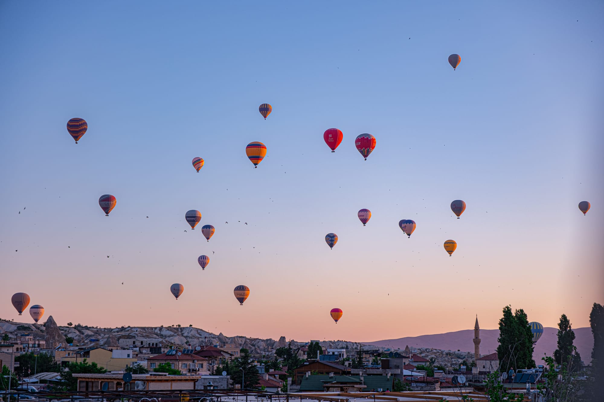 Dozens of colorful hot air balloons float above Cappadocia at sunrise, with fairy chimneys, stone houses, and a mosque minaret visible in the valley below against a pastel sky of pink and blue