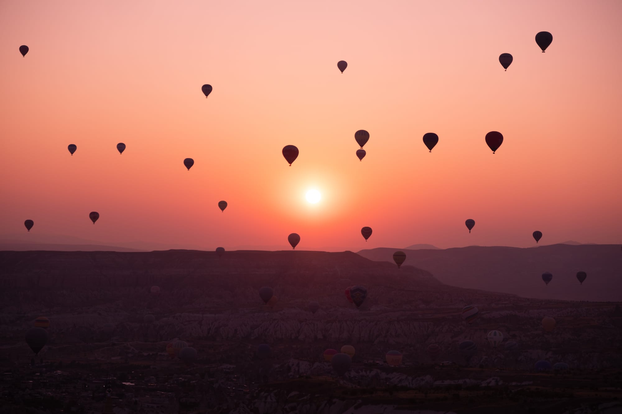 Dozens of hot air balloons float above Cappadocia at sunrise, silhouetted against a glowing orange and pink sky as the sun rises over the dark ridges and valleys below.