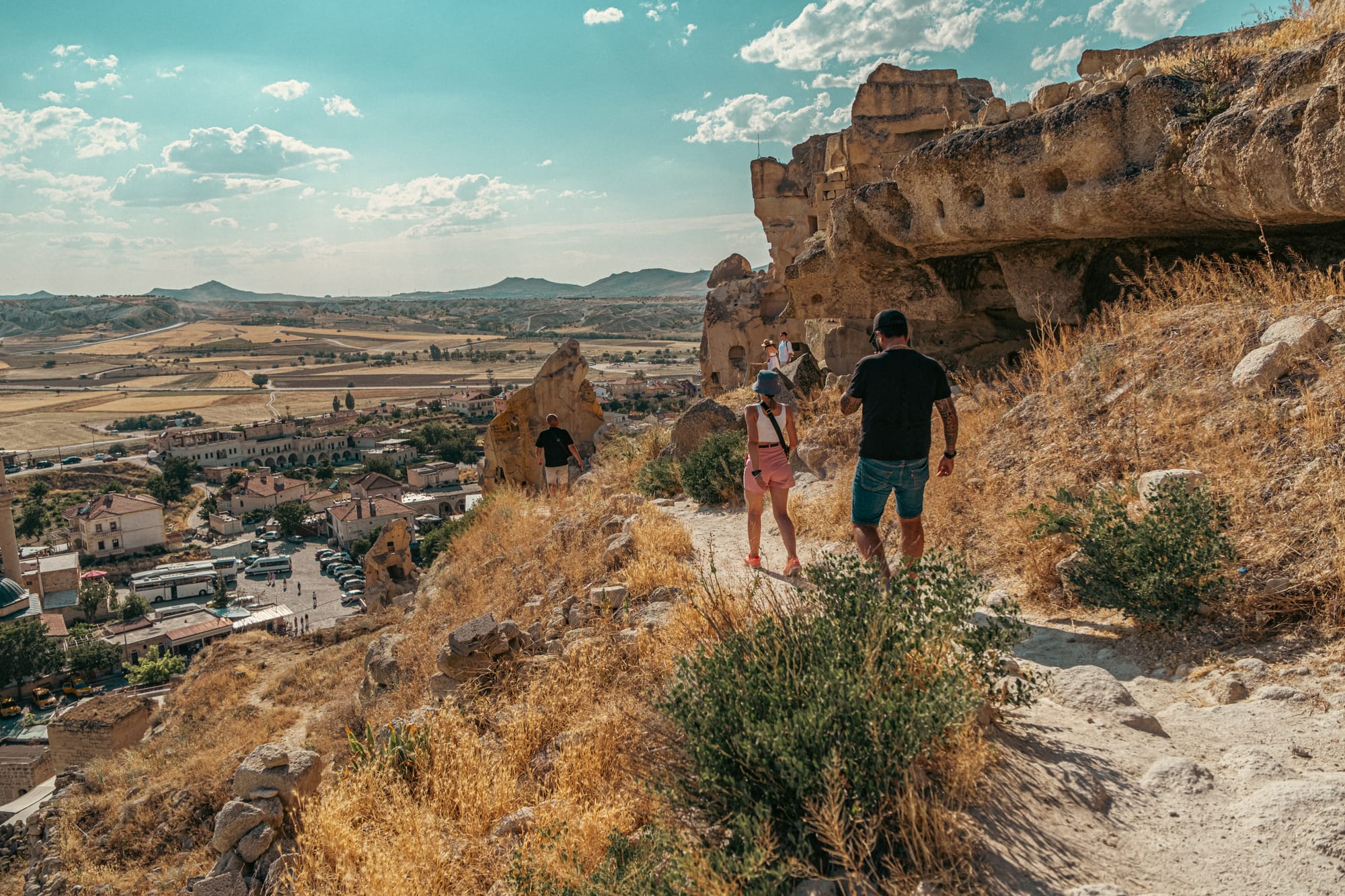 Visitors walking along the rocky trail to Çavuşin Castle with panoramic views of Cappadocia’s valleys and village below