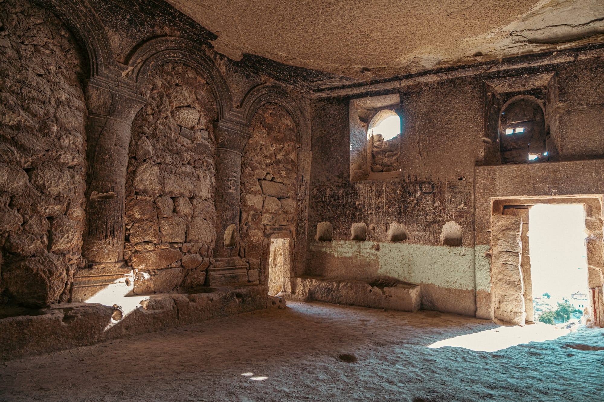 Interior of Çavuşin Castle’s ancient stone hall with arched niches, rough rock walls, and sunlight streaming through windows and doorways