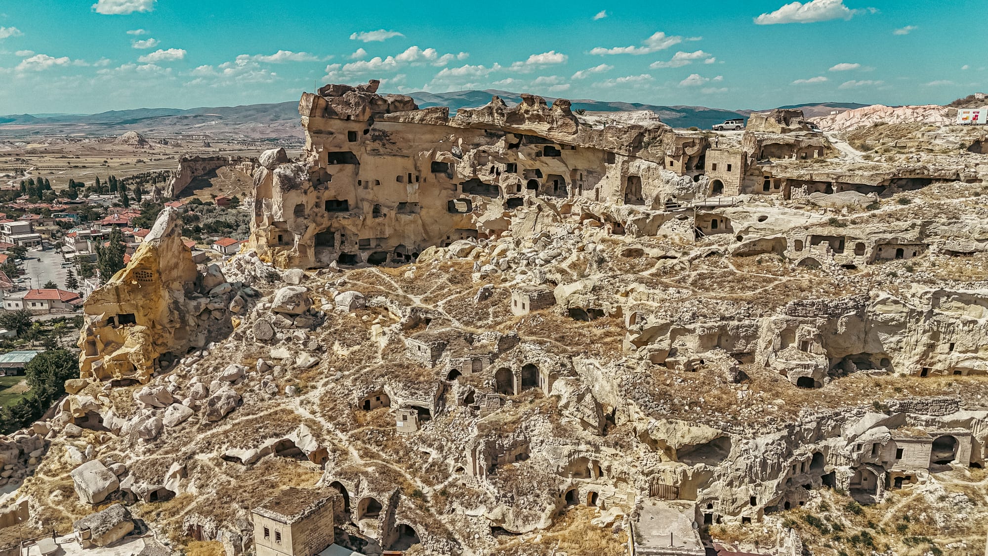 Aerial view of Çavuşin Castle in Cappadocia, Turkey, showcasing its sprawling network of ancient cave dwellings, carved rock facades, and surrounding village landscape under a bright blue sky with scattered clouds