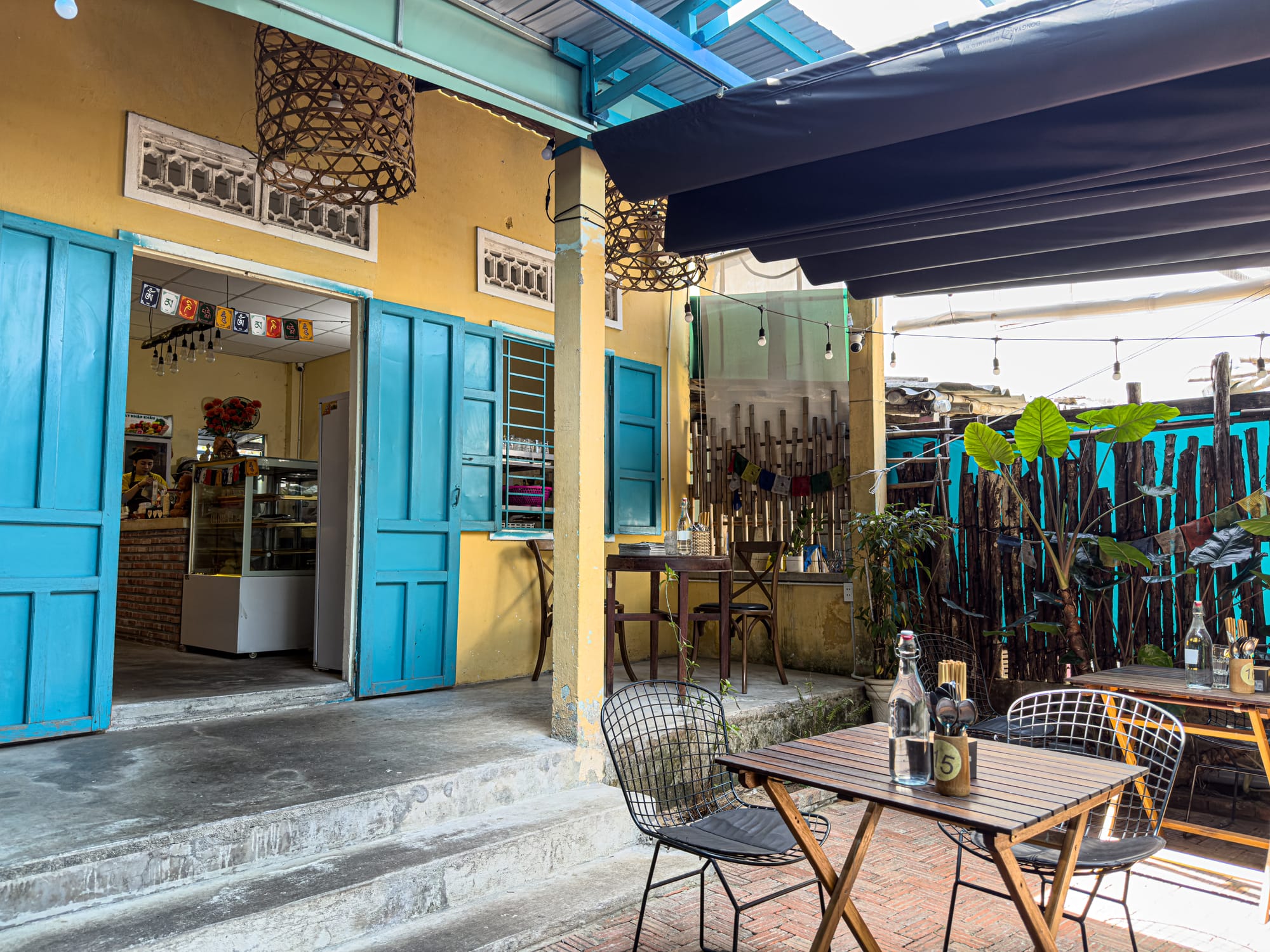 Outdoor courtyard at Chickpea Eatery in Hội An with yellow walls, turquoise shutters, wooden tables, black chairs, hanging lanterns, string lights, and plants creating a cozy atmosphere