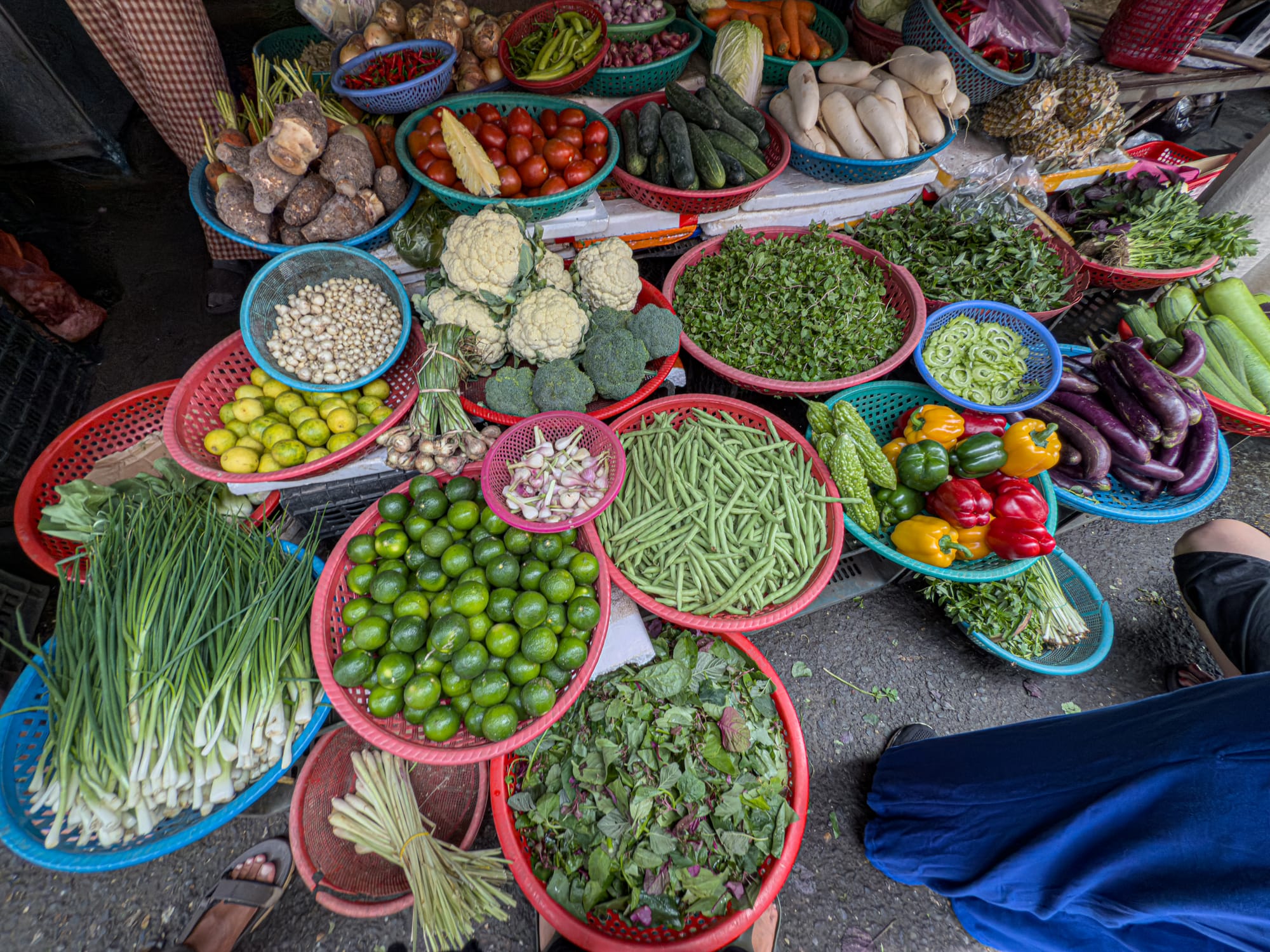 Fresh produce stall at a local market in Hội An with baskets of green beans, limes, scallions, broccoli, cauliflower, eggplant, bell peppers, cucumbers, tomatoes, herbs, and root vegetables used in Vietnamese vegan cooking classes
