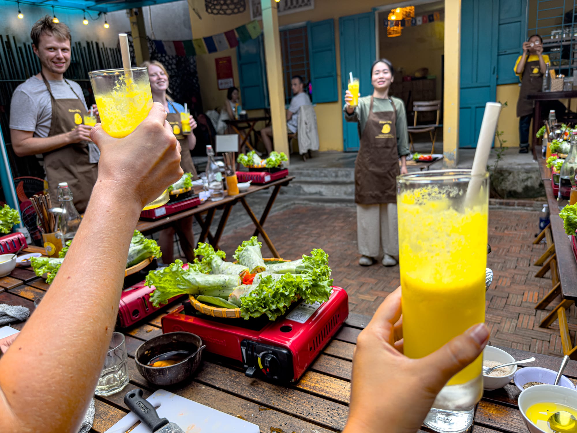 Participants at Chickpea Eatery's vegan cooking class in Hội An raising glasses of fresh pineapple juice for a toast around courtyard tables filled with spring rolls and cooking supplies