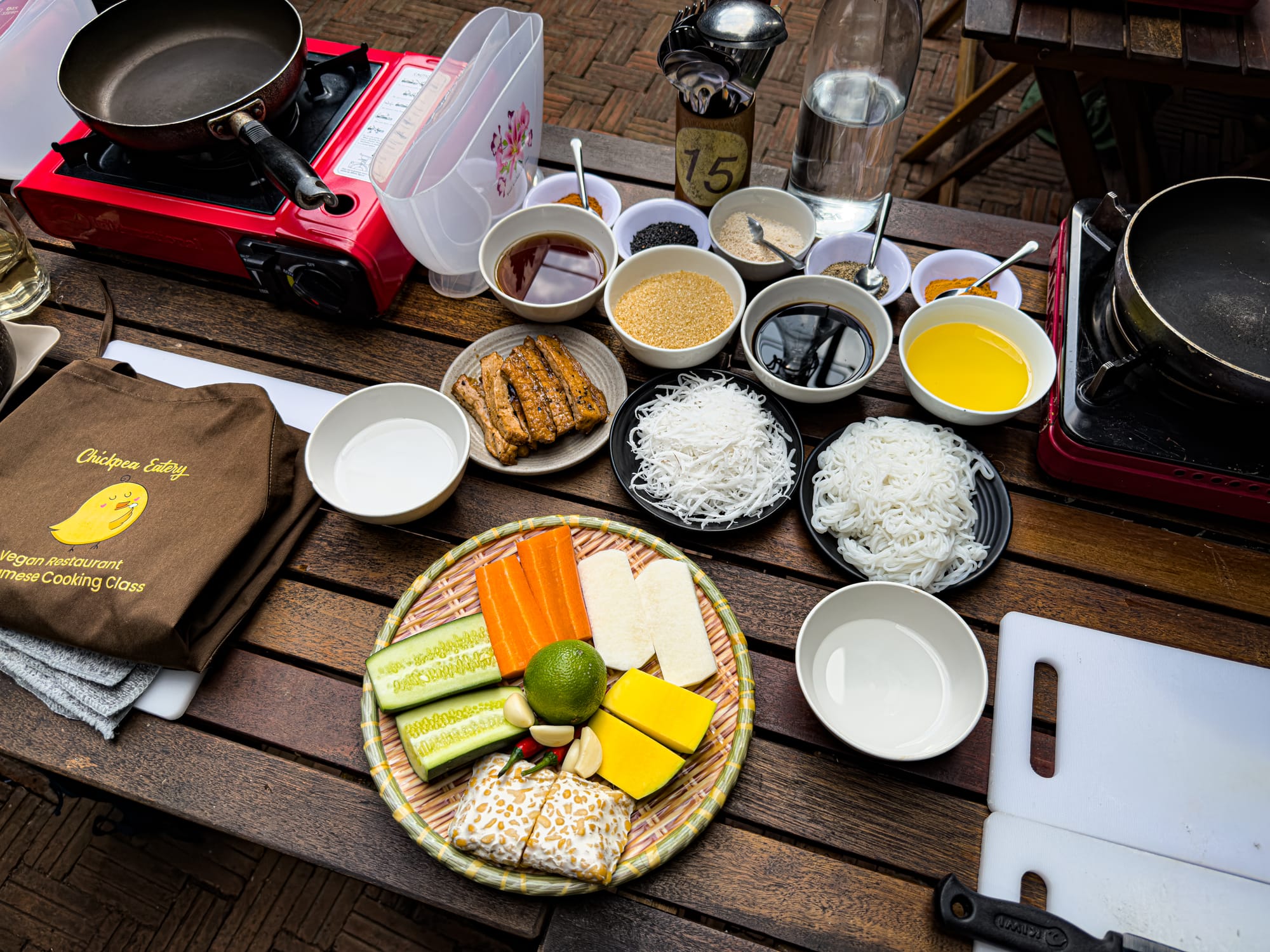 Table setup at Chickpea Eatery vegan cooking class in Hội An with gas burners, bowls of rice noodles, sauces, spices, marinated tofu skin, chickpea tempeh, fresh vegetables, and cutting boards ready for preparing Vietnamese dishes