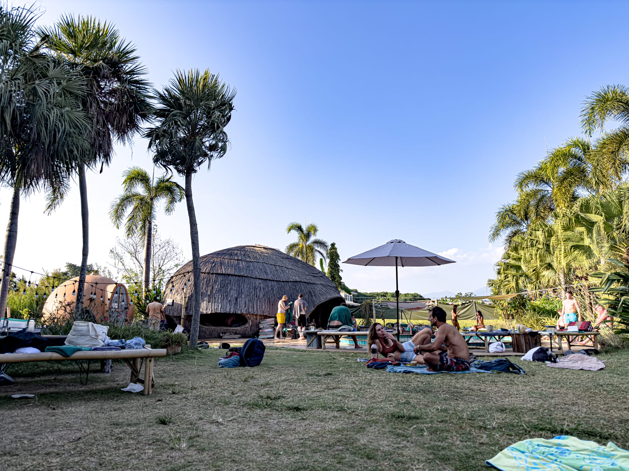Outdoor gathering area at Cocoon in Chiang Mai with people lounging on mats and benches near dome-shaped thatched and clay structures, framed by palm trees and tropical greenery
