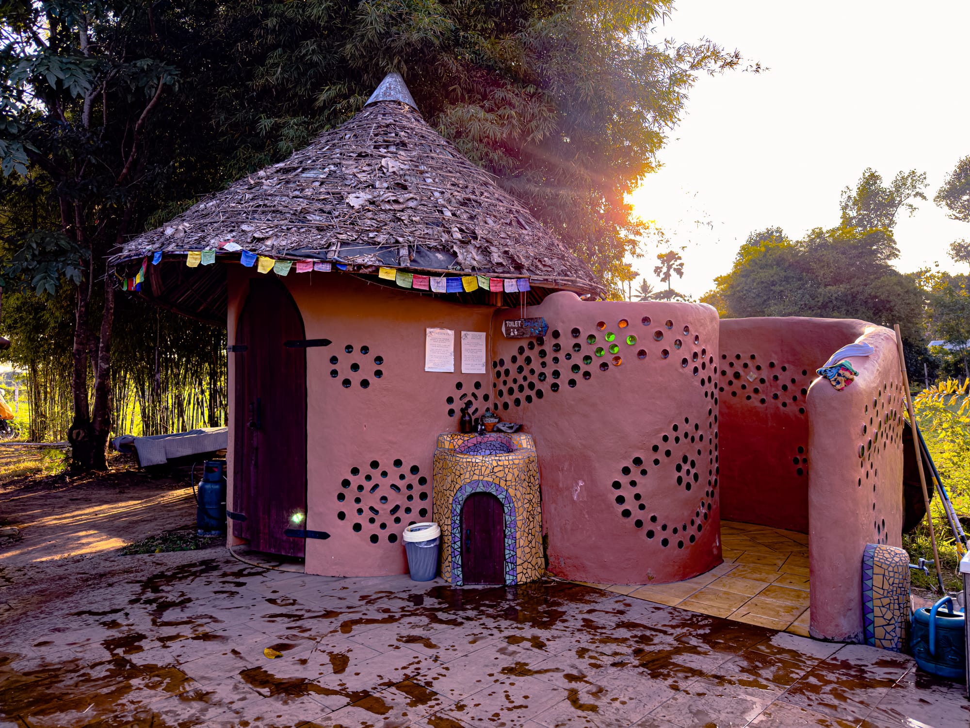 Round clay-built structure at Cocoon Chiang Mai with a thatched roof, prayer flags, and mosaic details, designed in organic curves with natural materials, blending into the surrounding bamboo forest