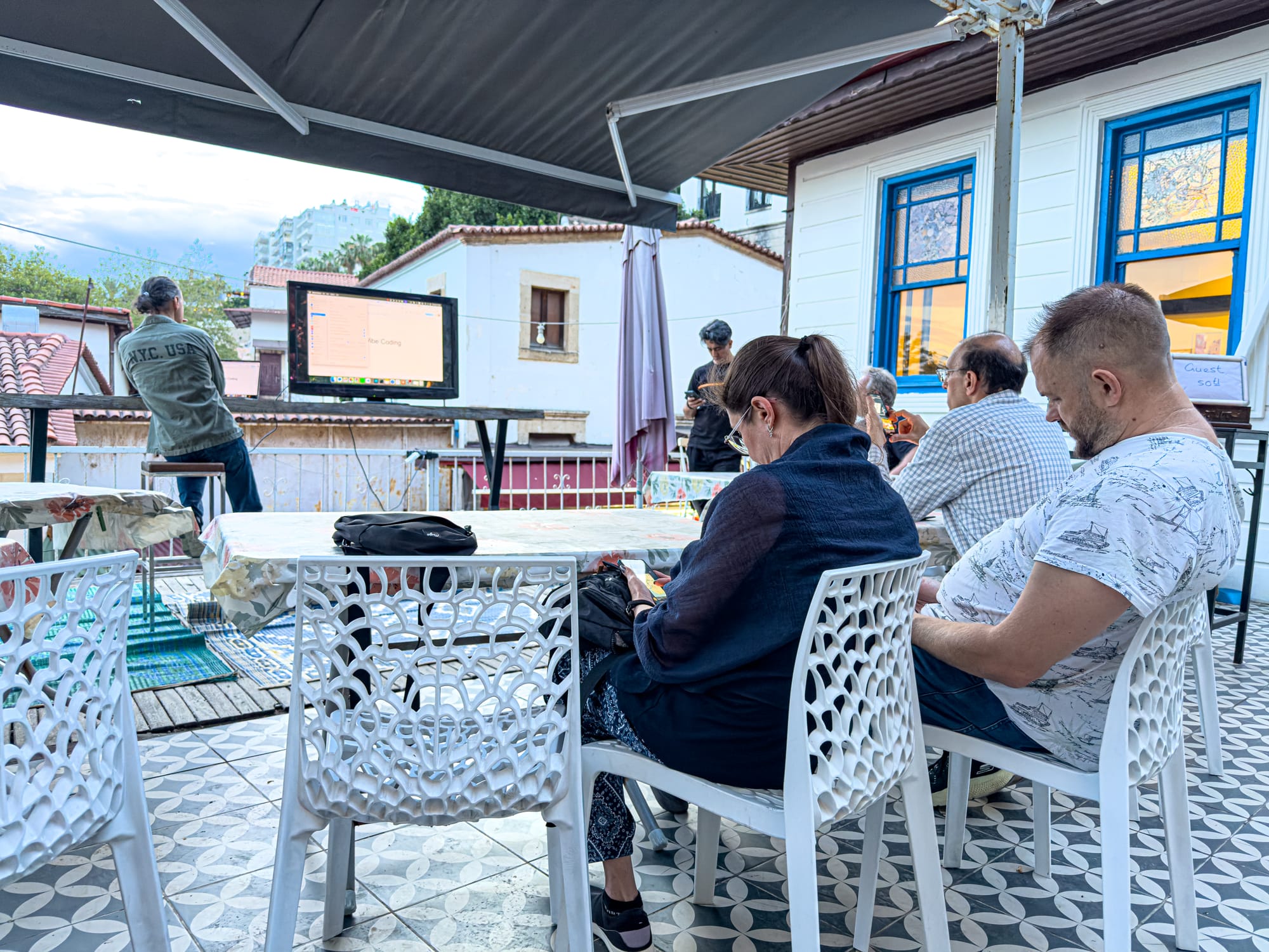 People seated at outdoor tables under a canopy at Coworking Antalya, listening to a presenter speaking next to a large screen