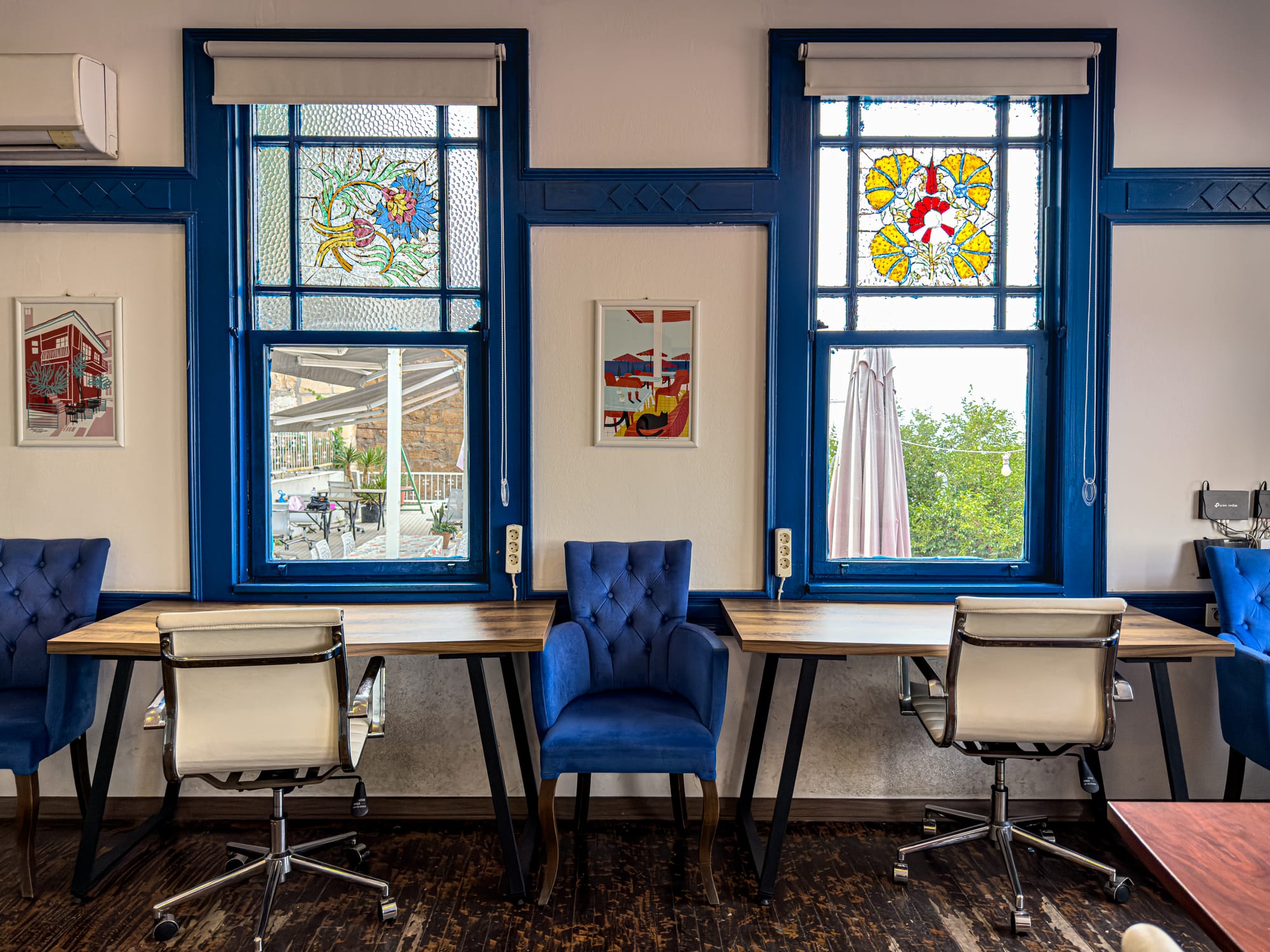 Two desks with office chairs sit in front of blue-framed stained-glass windows at Coworking Antalya, offering a bright and stylish workspace