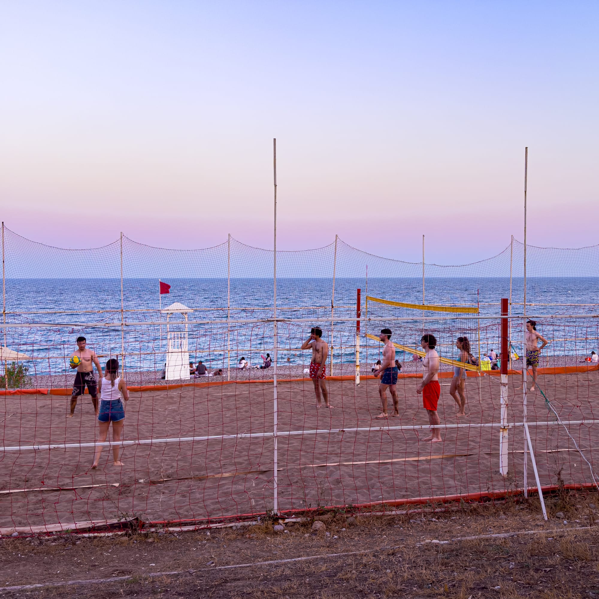 Seven people playing beach volleyball on a sand court beside the sea, with a lifeguard tower and swimmers in the background under a pastel evening sky