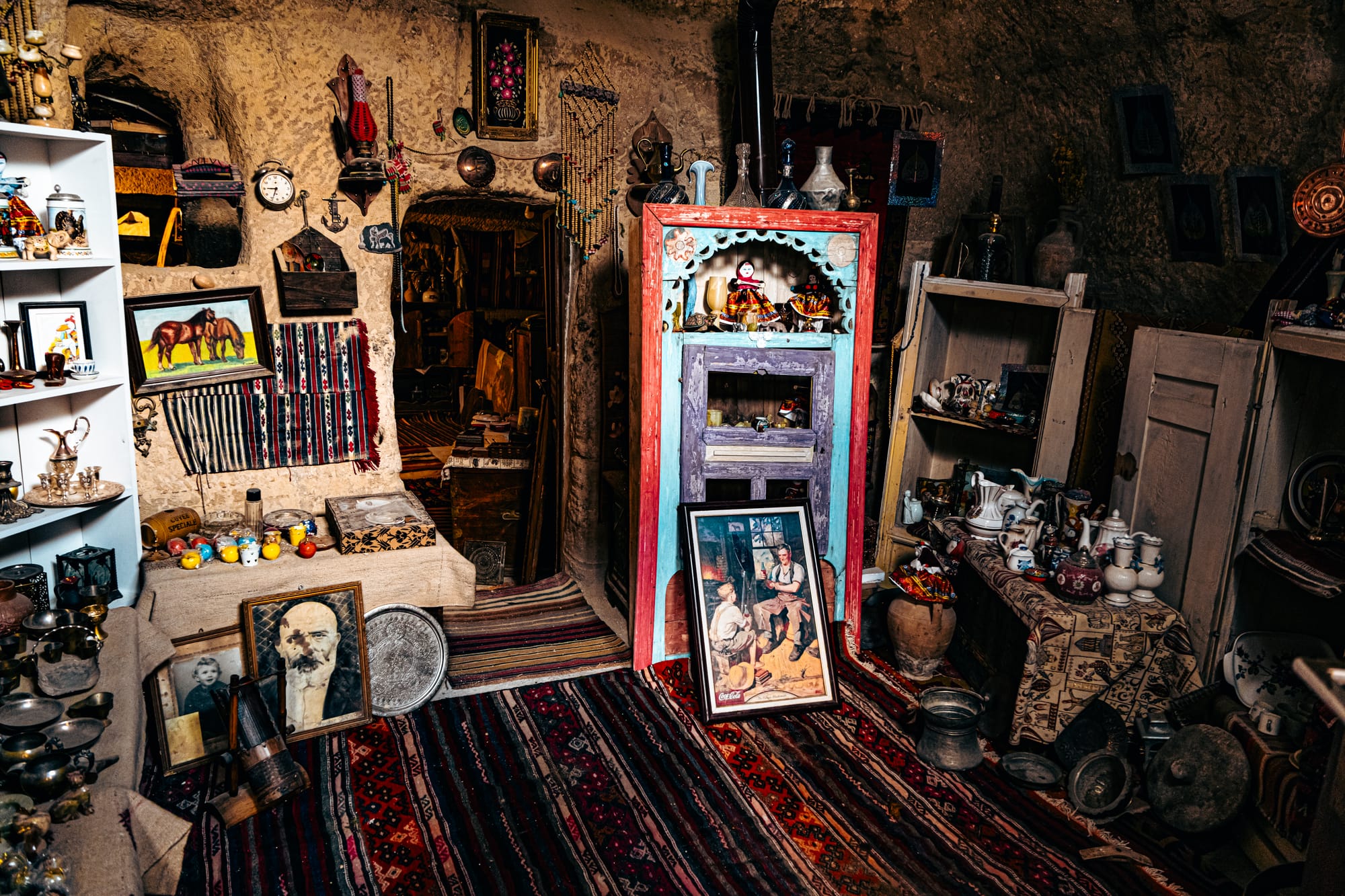 Interior of Cappadocia Cultural Antique Collection cave room with colorful rugs, vintage furniture, and shelves filled with antique pottery, framed artwork, and traditional Anatolian artifacts