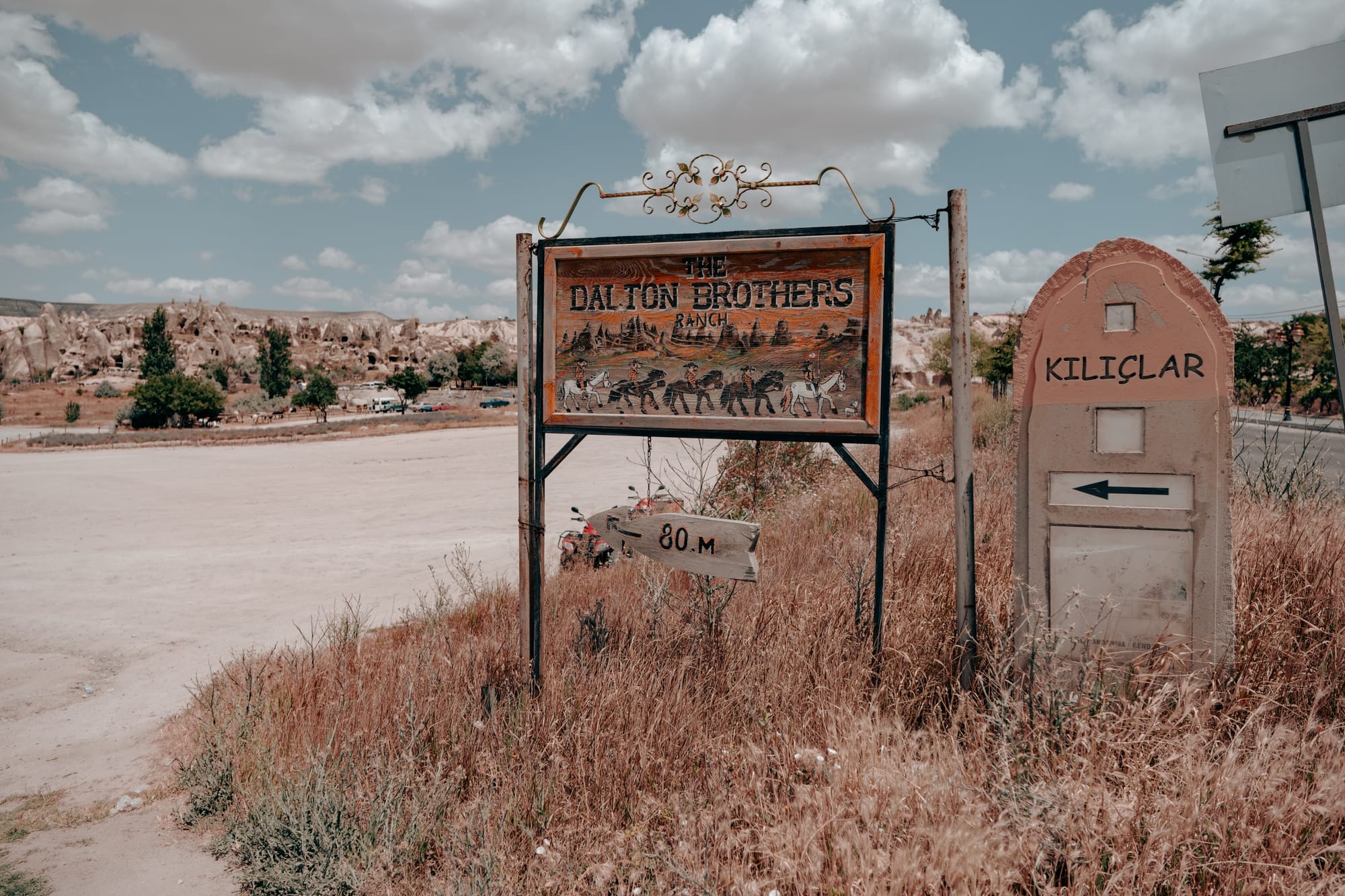 Entrance signage for Dalton Brothers Ranch, featuring a rustic illustrated wooden board and an adjacent stone marker reading “Kılıçlar,” surrounded by dry grass and backed by Cappadocia’s rocky landscape under a partly cloudy sky