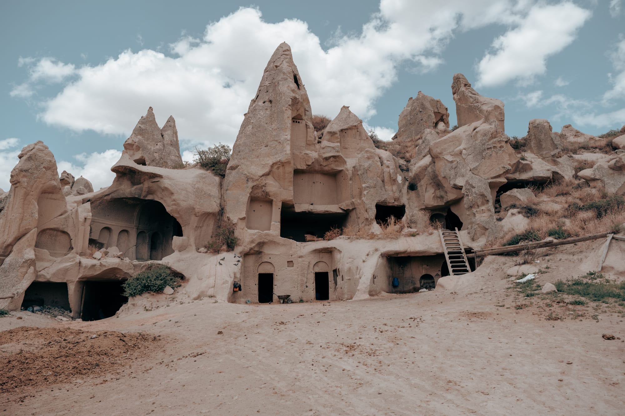 Ancient rock-carved dwellings with arched entrances and wooden ladders built into towering cliffs at Dalton Brothers Ranch in Cappadocia, Turkey, showcasing the region’s unique cave architecture under a partly cloudy sky