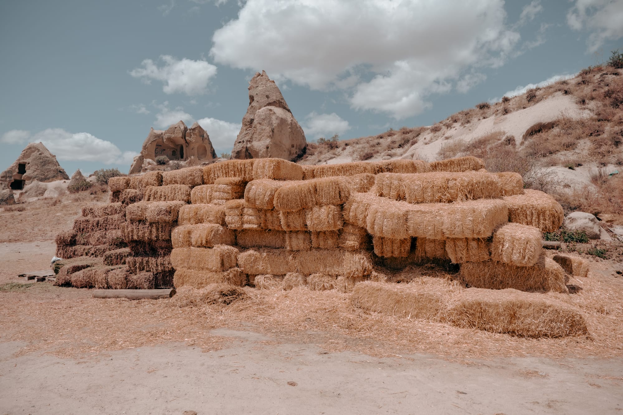 Large stack of golden hay bales at Dalton Brothers Ranch in Cappadocia, Turkey, with distinctive fairy chimney rock formations and ancient cave dwellings in the background under a partly cloudy sky