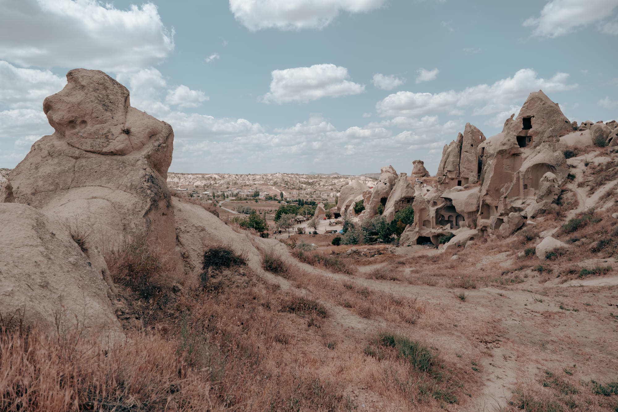 Panoramic view from Dalton Brothers Ranch in Cappadocia, Turkey, featuring weathered rock formations, ancient cave dwellings, and a vast valley landscape with scattered greenery and distant settlements under a partly cloudy sky