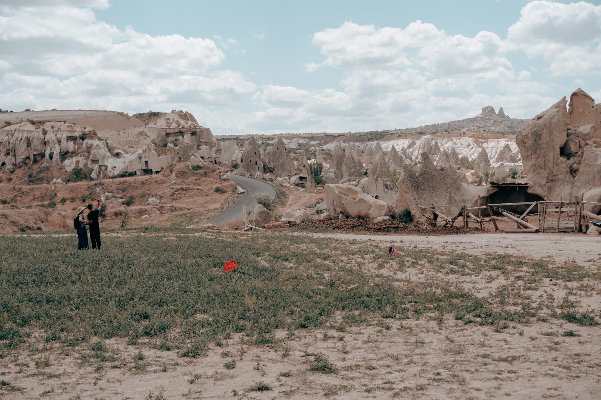 Wide view of Dalton Brothers Ranch area in Cappadocia, Turkey, showing visitors standing in a grassy field with a winding road leading through dramatic fairy chimney rock formations and ancient cave dwellings under a partly cloudy sky