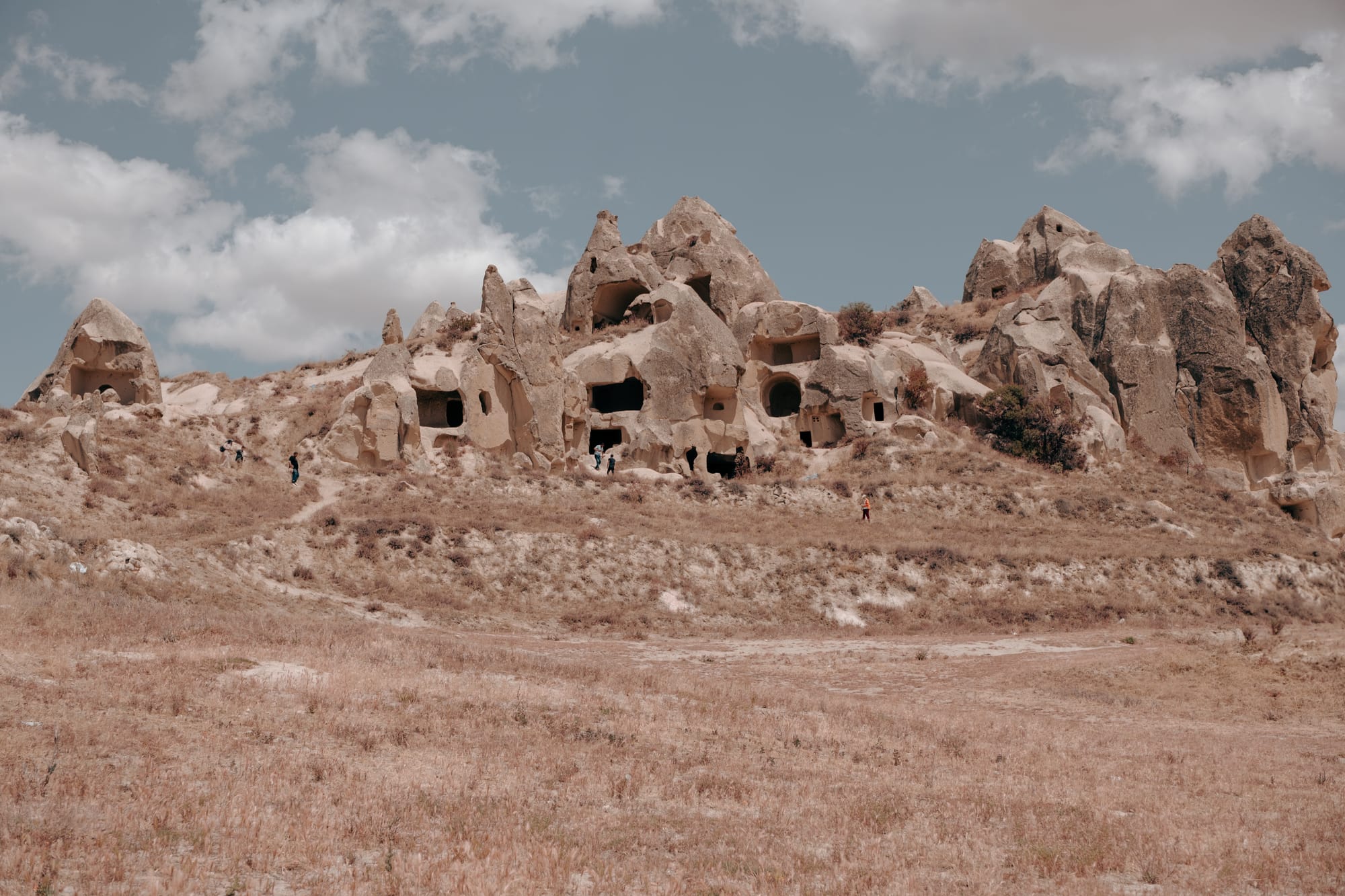 Expansive view of ancient cave dwellings carved into rocky cliffs at Dalton Brothers Ranch in Cappadocia, Turkey, with visitors exploring the site under a partly cloudy sky