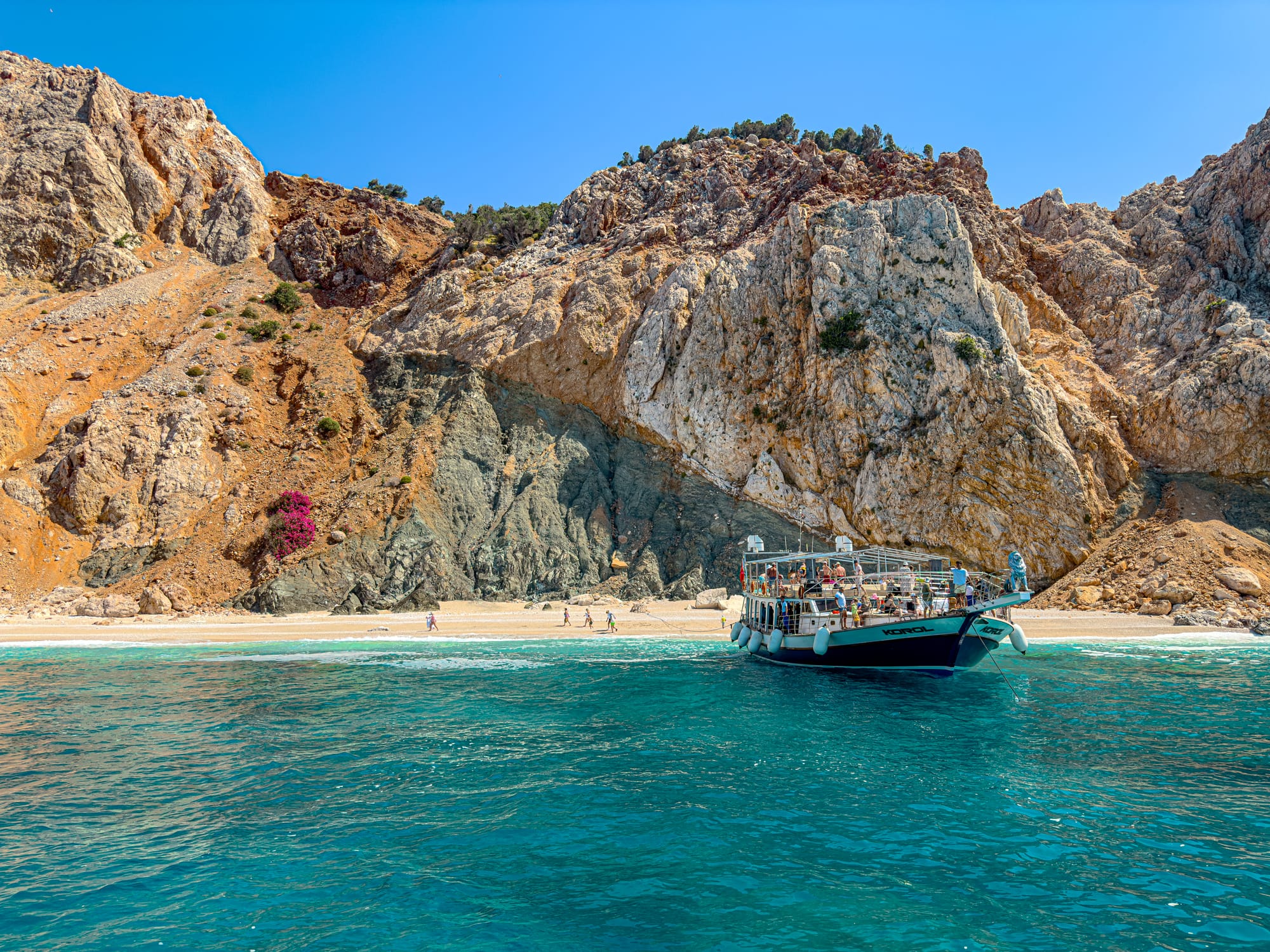 A small tour boat anchored at a remote beach near Suluada Island, backed by dramatic red and grey cliffs