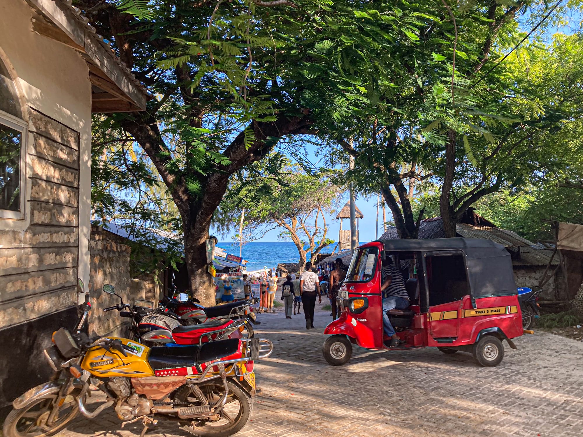 Motorbikes and a red tuk-tuk line a shaded street in Diani as people walk toward the Indian Ocean, visible at the end of the road beneath a canopy of green trees