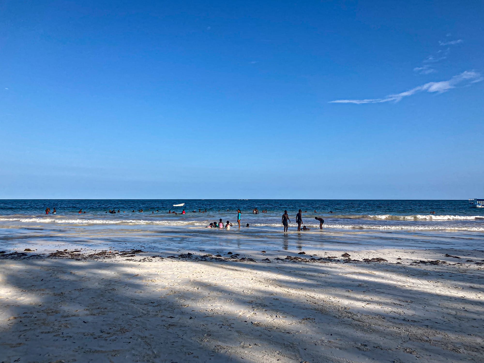People swim, play, and relax along the shoreline at Diani Beach, with gentle waves rolling in under a wide blue sky and the white sand stretching into the foreground