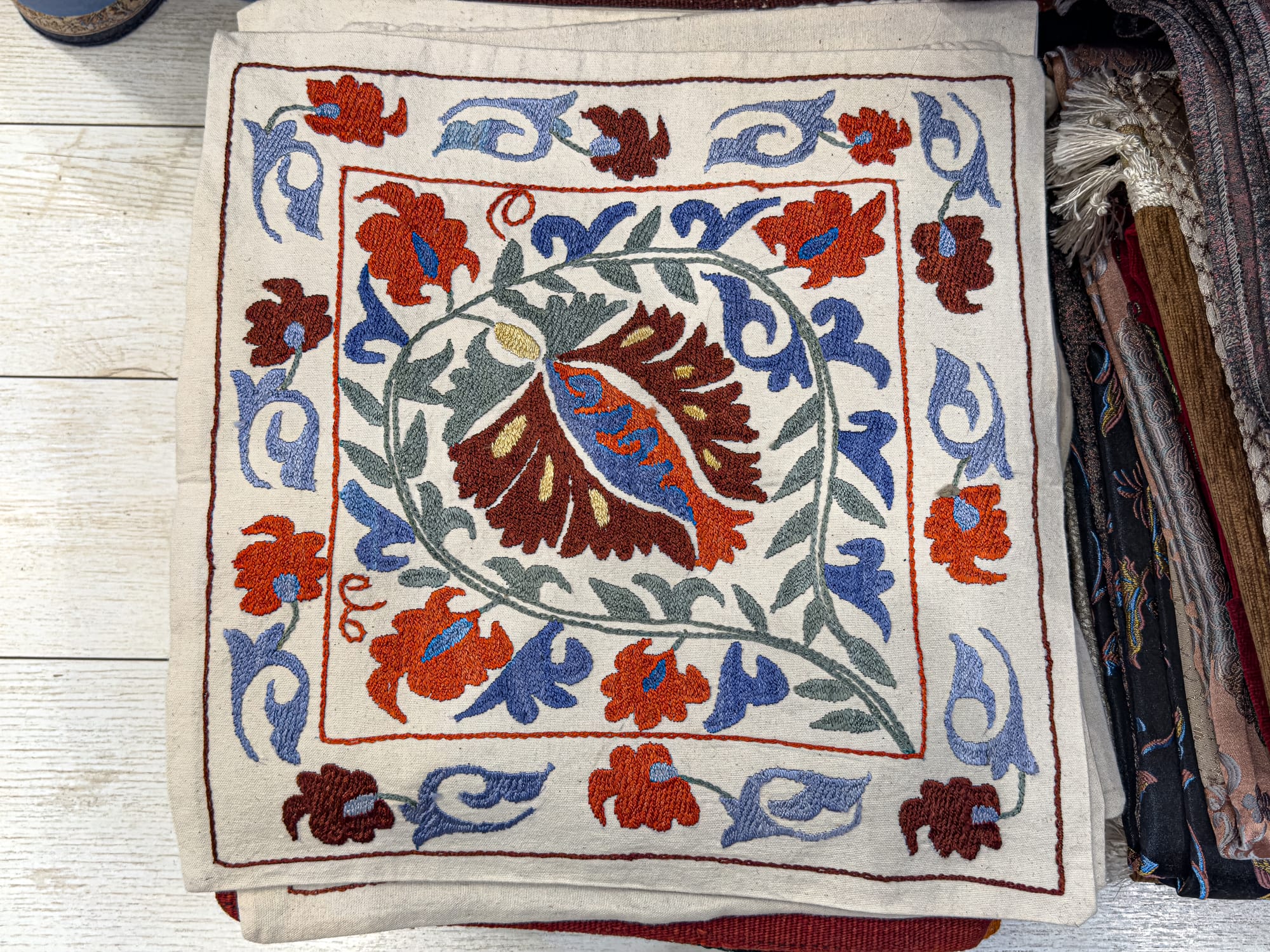Close-up of a square embroidered textile featuring a central floral design with red, blue, and green leaves, bordered by repeating flower patterns on a cream-colored fabric