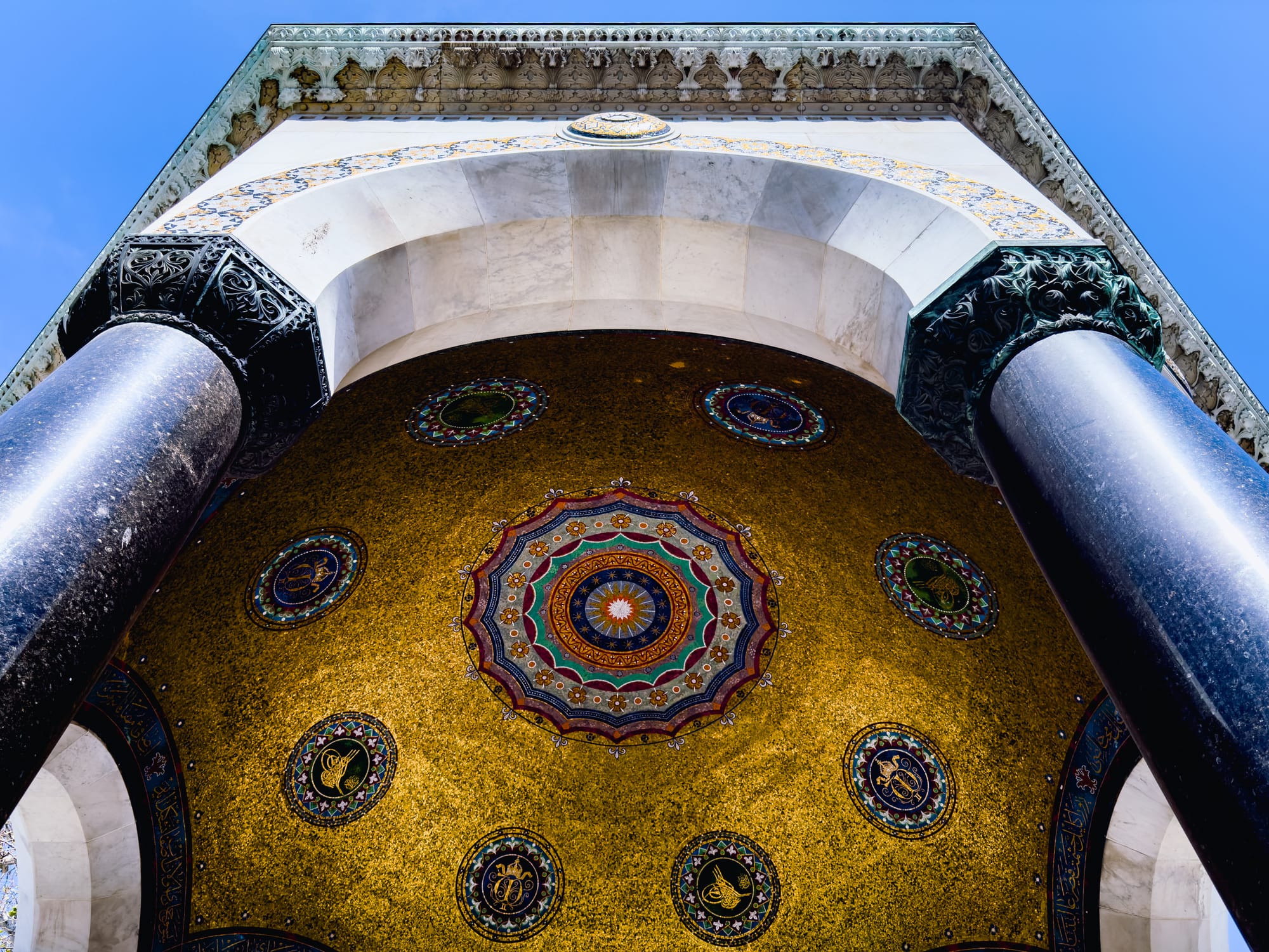 Upward view of the German Fountain’s domed ceiling in Istanbul, adorned with intricate golden mosaics and colorful geometric patterns, framed by black marble columns against a clear blue sky