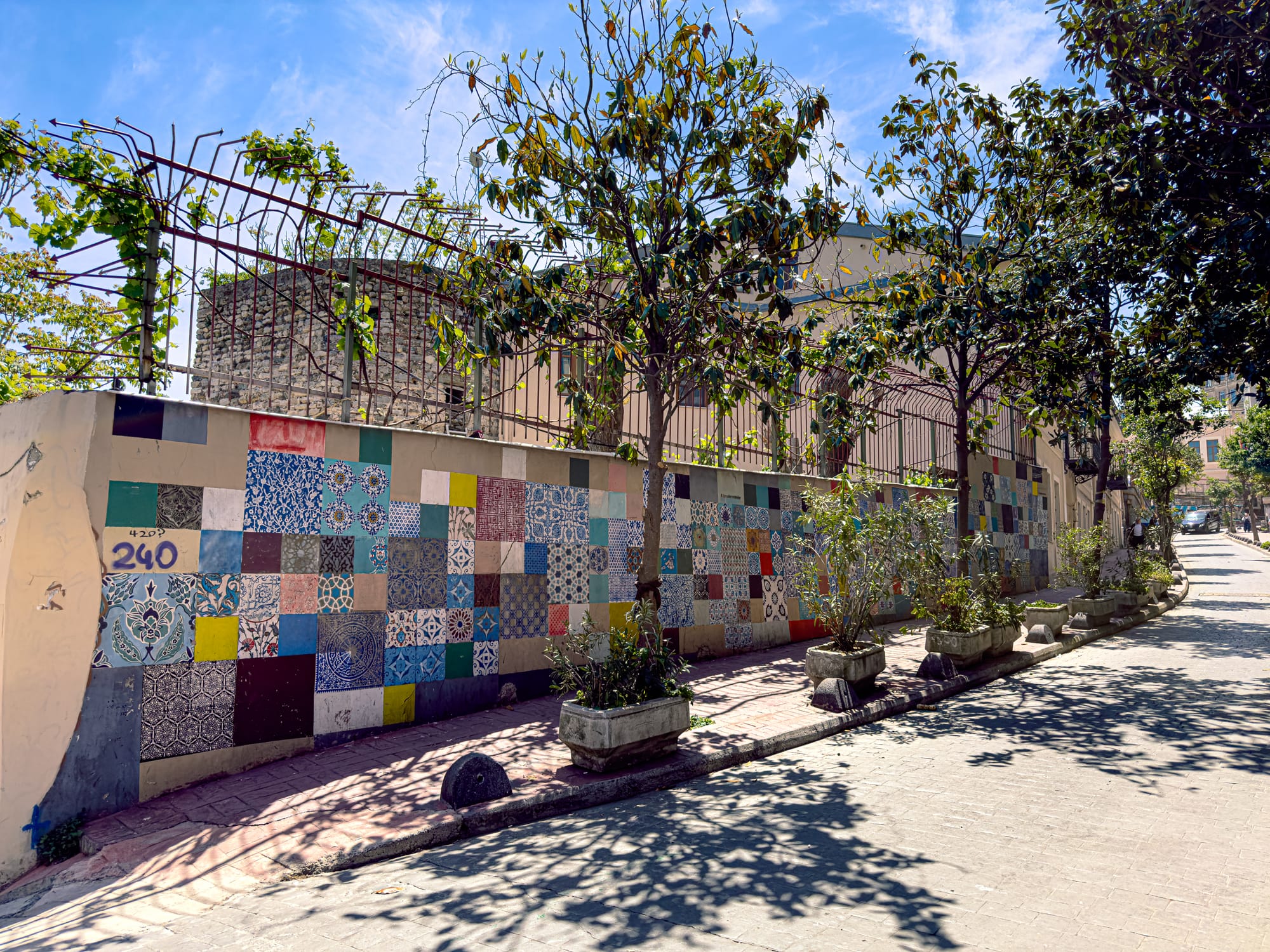 A sunlit street in Istanbul featuring a wall decorated with an eclectic patchwork of colorful patterned tiles, lined with potted plants and shaded by leafy trees