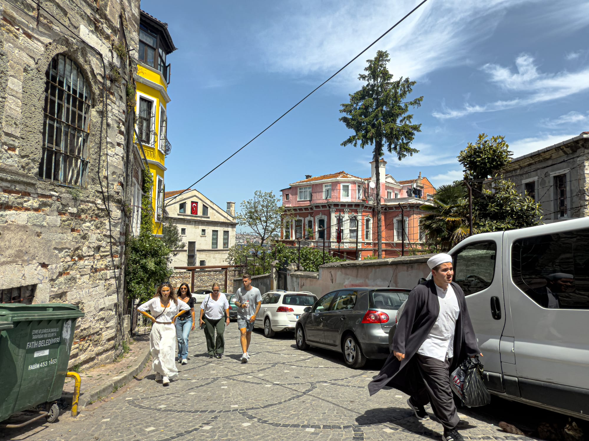 A lively Istanbul street scene showing people walking along a cobblestone road, framed by weathered stone walls, a bright yellow building, and pastel-colored houses under a blue sky with wispy clouds