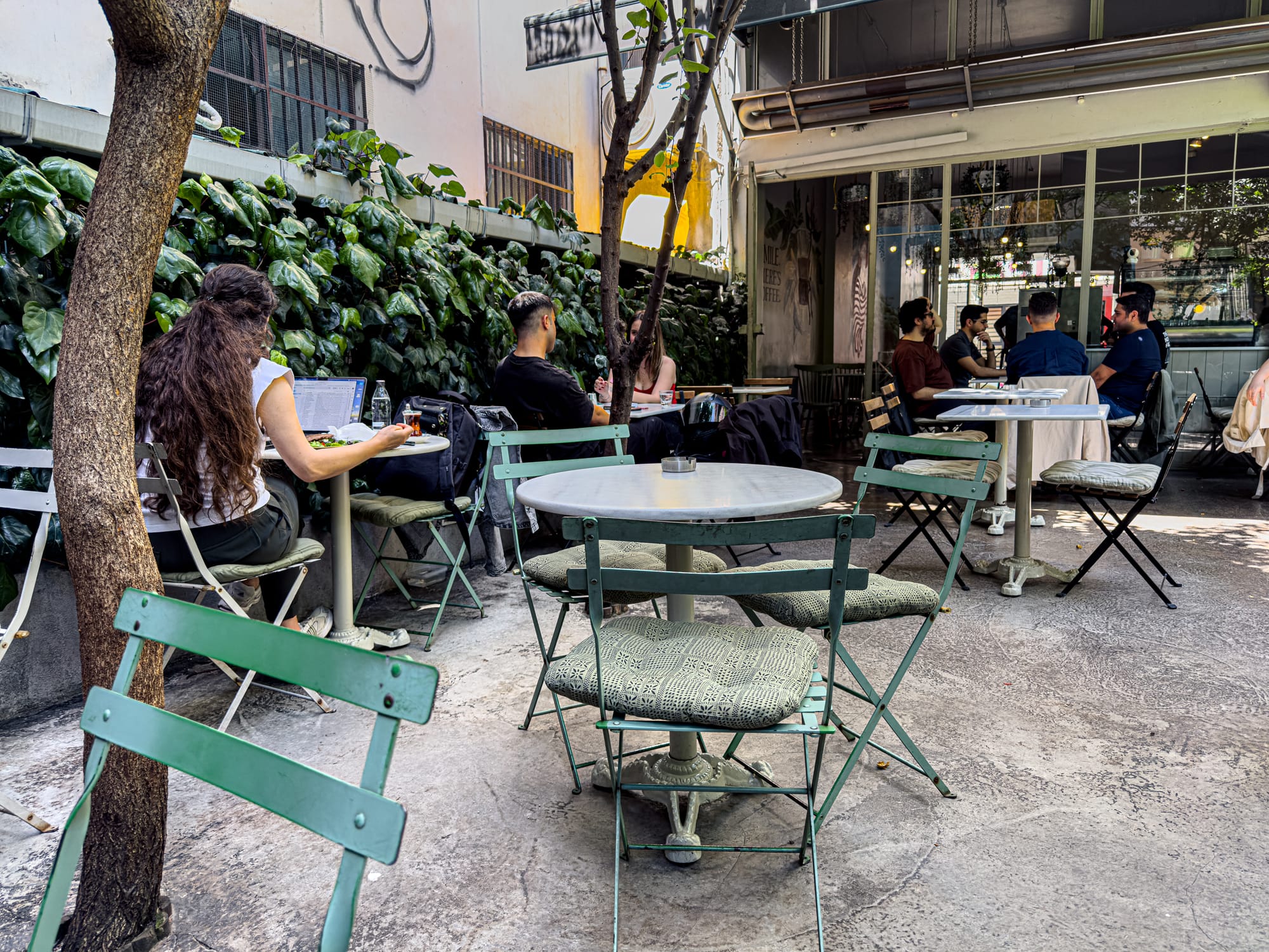 Outdoor café in Istanbul with green metal chairs, round tables, and leafy wall backdrop, where people are working on laptops and chatting in the shaded courtyard