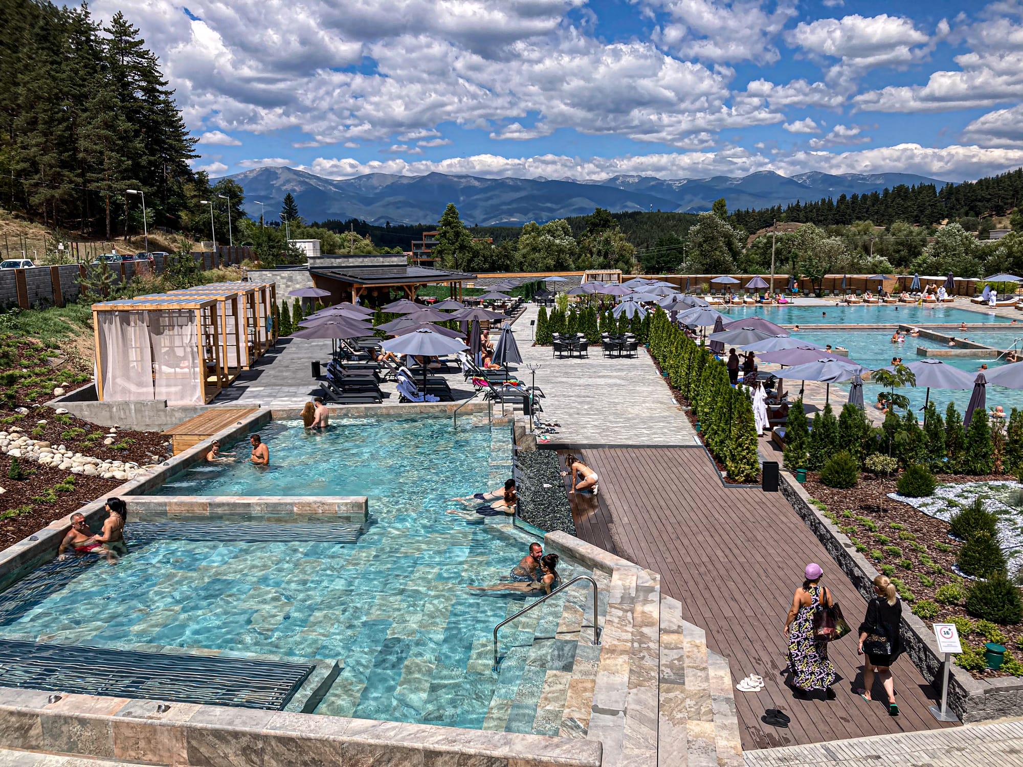 View of the outdoor mineral pools at Pulse Therme in Banya, Bulgaria, with people relaxing in the steaming turquoise water, lounge chairs shaded by umbrellas, and the Pirin Mountains visible in the distance under a partly cloudy sky