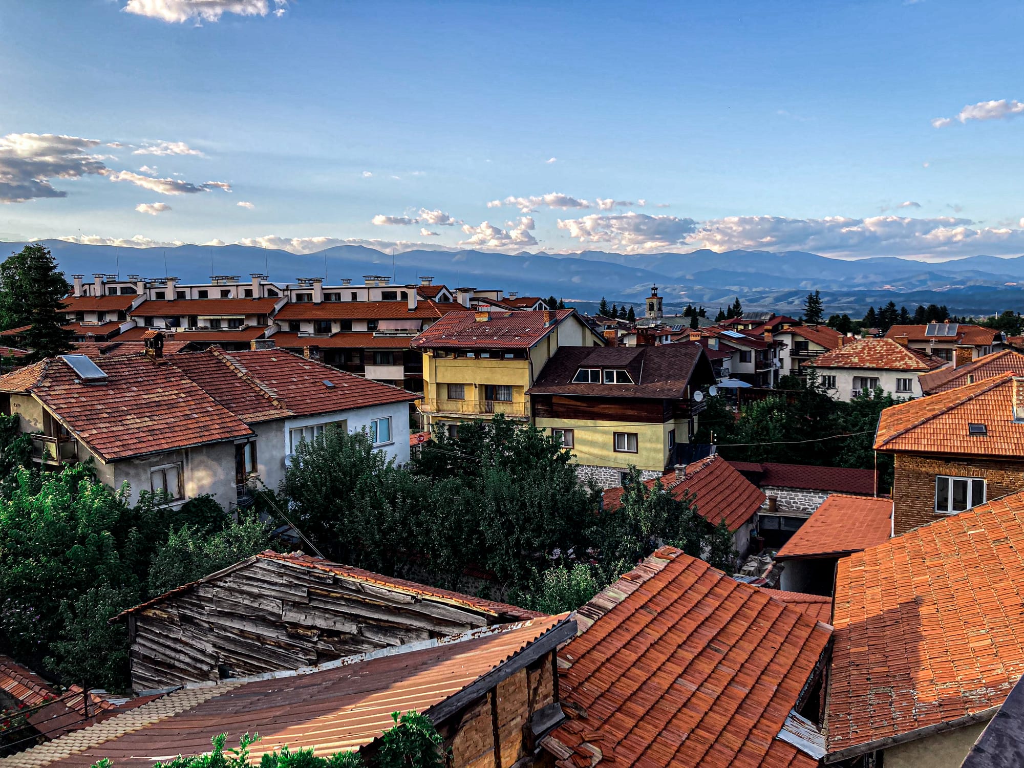 View over Bansko’s old town rooftops with red tiles and a mix of colorful houses, framed by the Pirin Mountains and a sky scattered with clouds at sunset